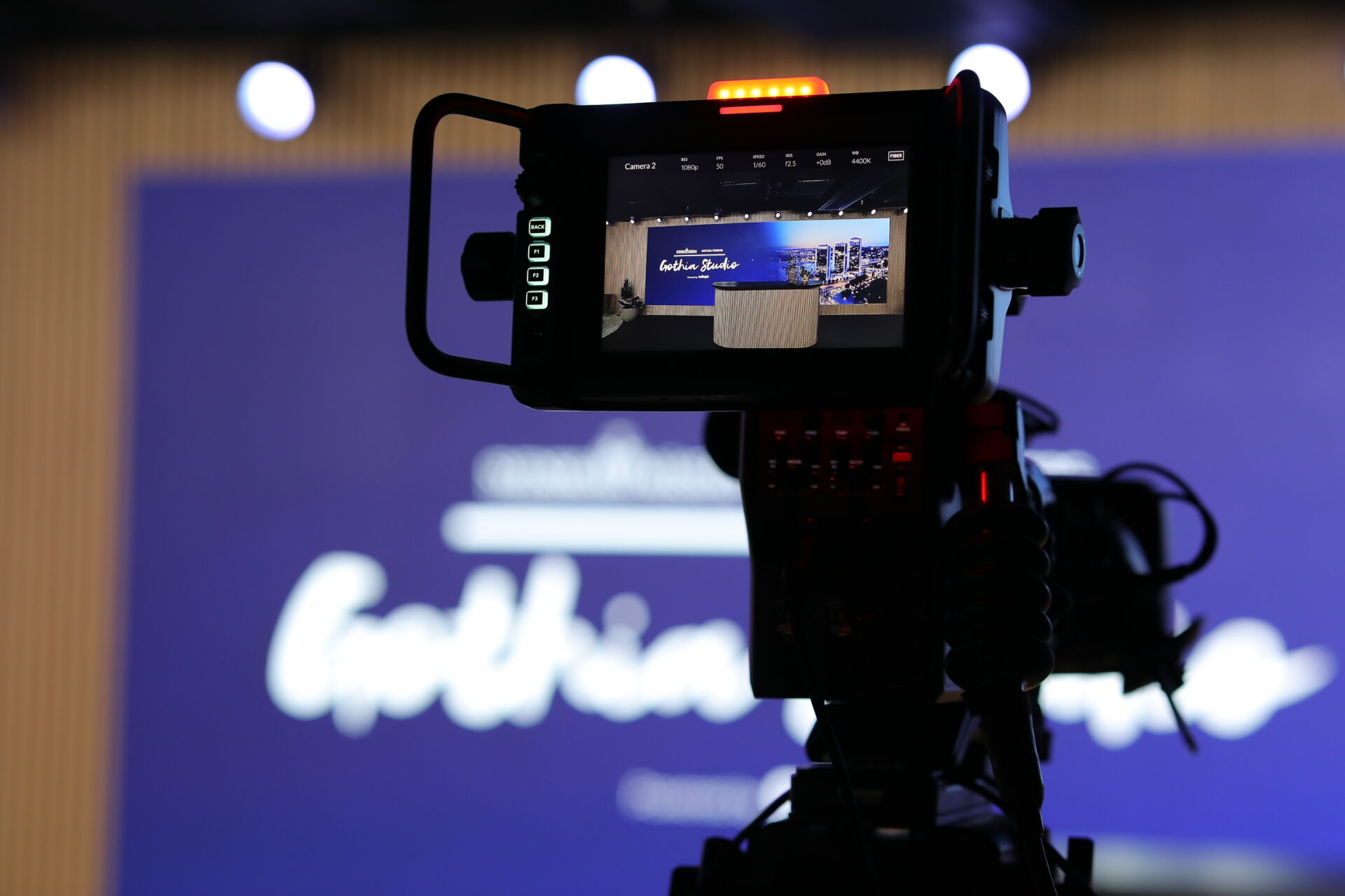 A close-up of a video camera recording an event, with the camera’s screen displaying a sharp view of the stage, while the background appears blurred and features blue and white colors.