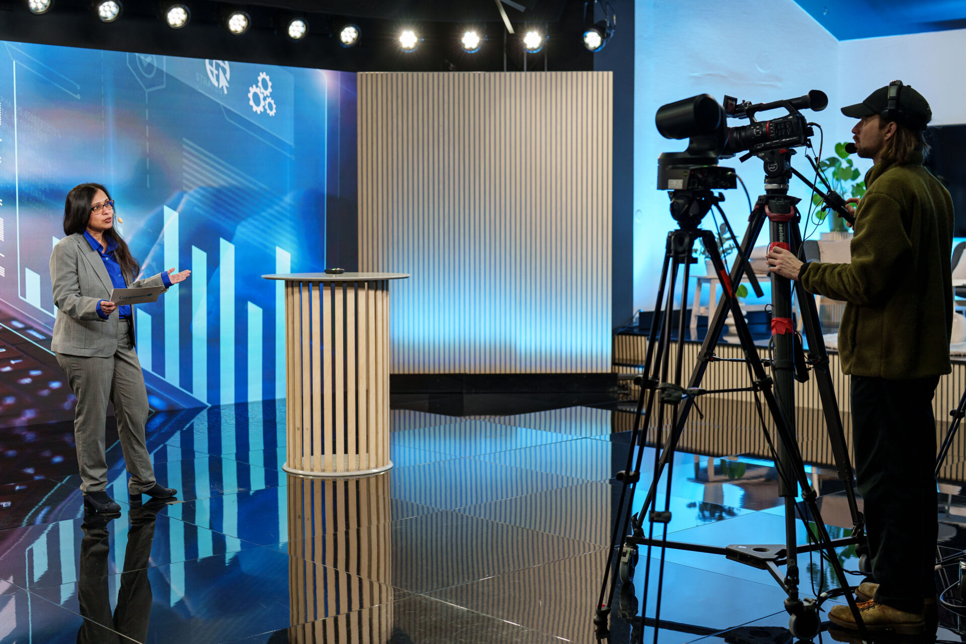 A woman in a gray suit presents on a modern TV studio set with blue lighting and a digital graph display, while a camera operator films her with professional video equipment.