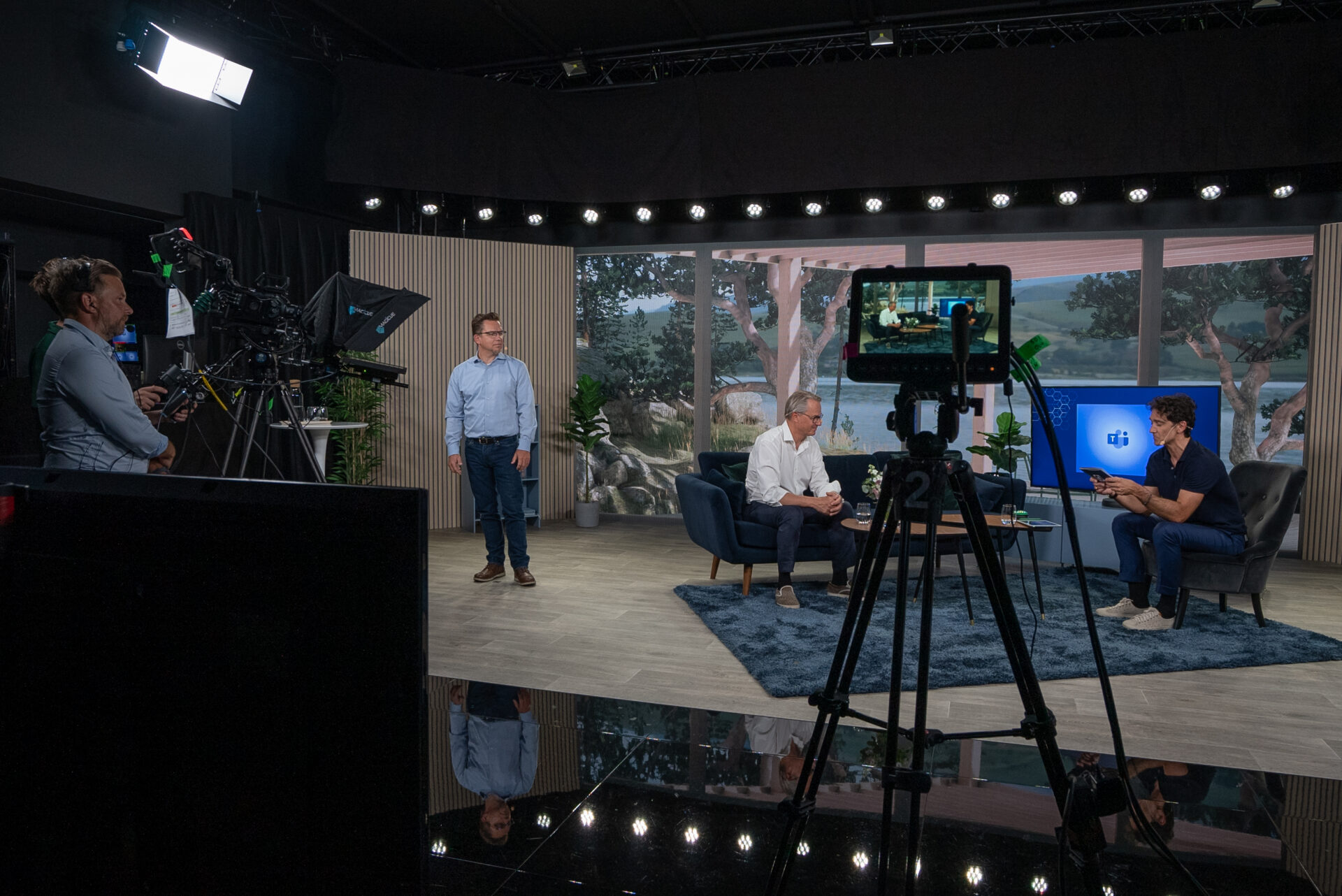A camera crew films two men having a conversation on a studio set with large windows, plants, and a monitor displaying a logo. Lighting equipment and cameras are visible, with crew members working in the background.