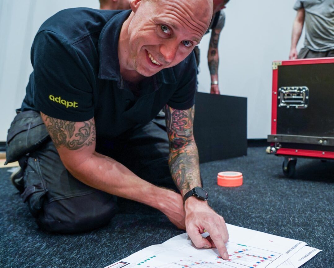 A smiling man kneels on the floor, pointing at a chart on paper. Behind him are equipment cases and two people standing, partially out of frame. The setting appears to be a work or event setup environment.