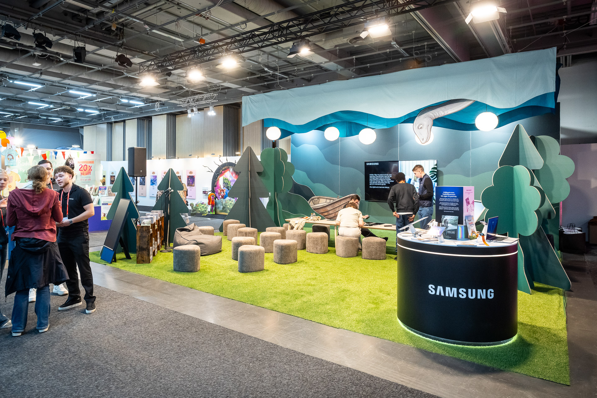 A Samsung booth at an indoor event features artificial grass, round cushioned seats, tree decorations, and a blue backdrop with mountains and clouds. People are interacting near the booth and looking at electronic displays.