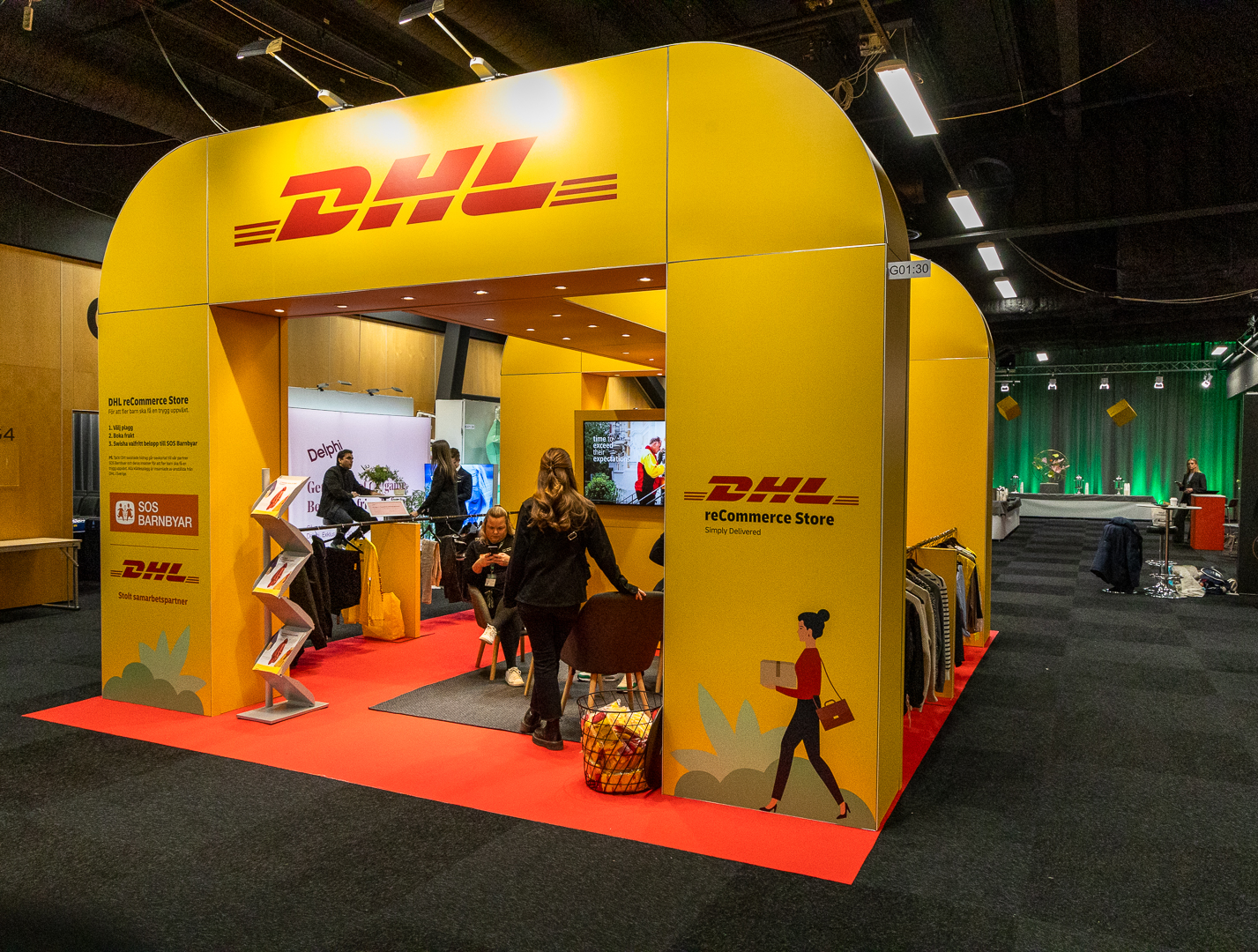 A bright yellow DHL booth at an indoor expo, with the DHL logo displayed prominently. People are interacting inside the booth, which features shelves, promotional items, and clothing displays on a red carpet.