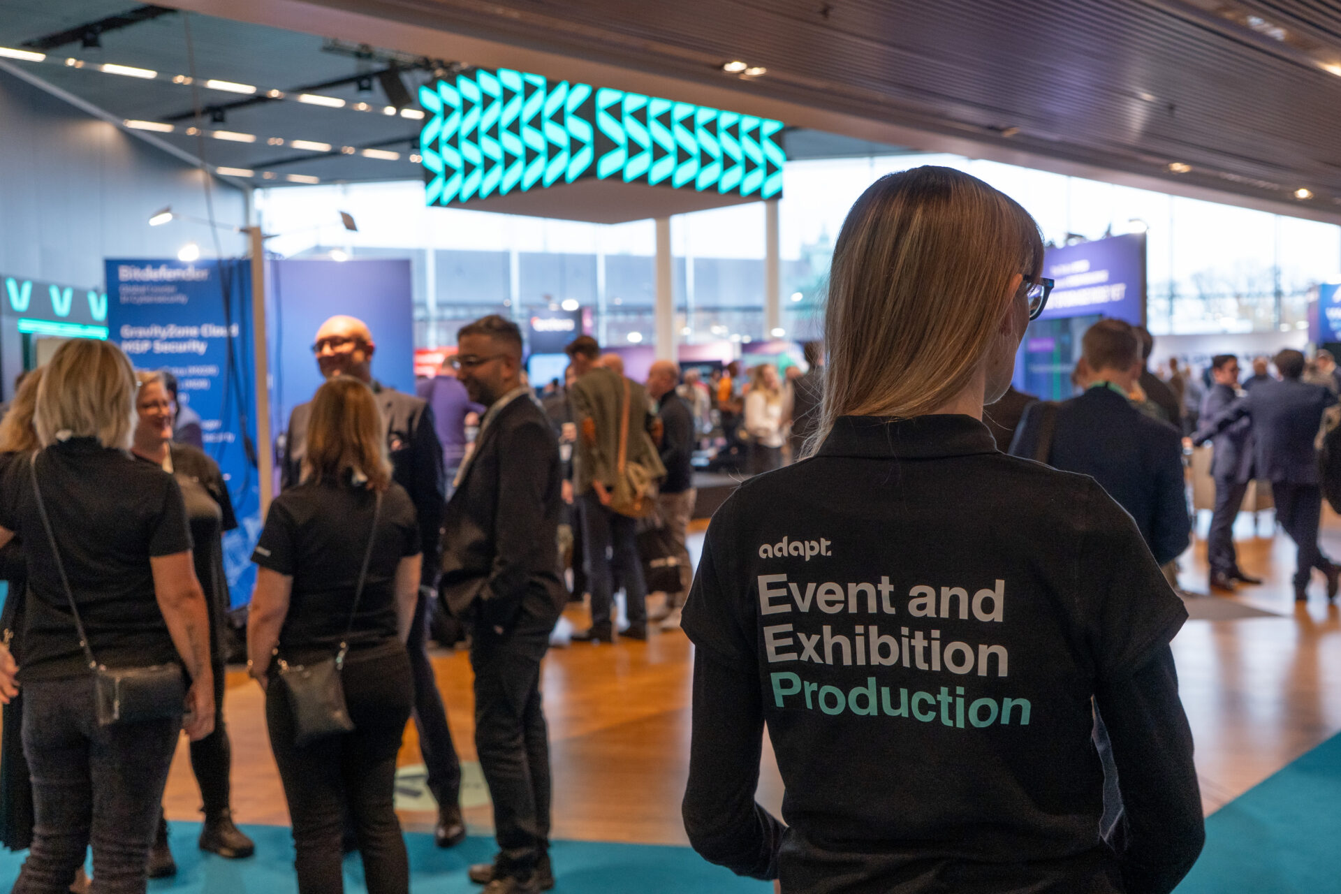 A person wearing a shirt labeled Event and Exhibition Production stands in a busy conference hall filled with people networking and talking near exhibition booths.