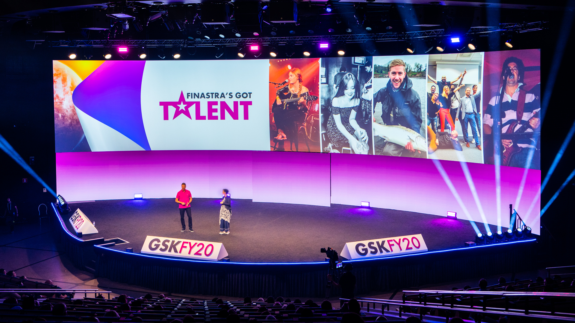 Two people stand on a stage in front of a large screen displaying Finastras Got Talent and photos of various performers. The stage is lit with purple and blue lights, and the audience seating is visible in the foreground.