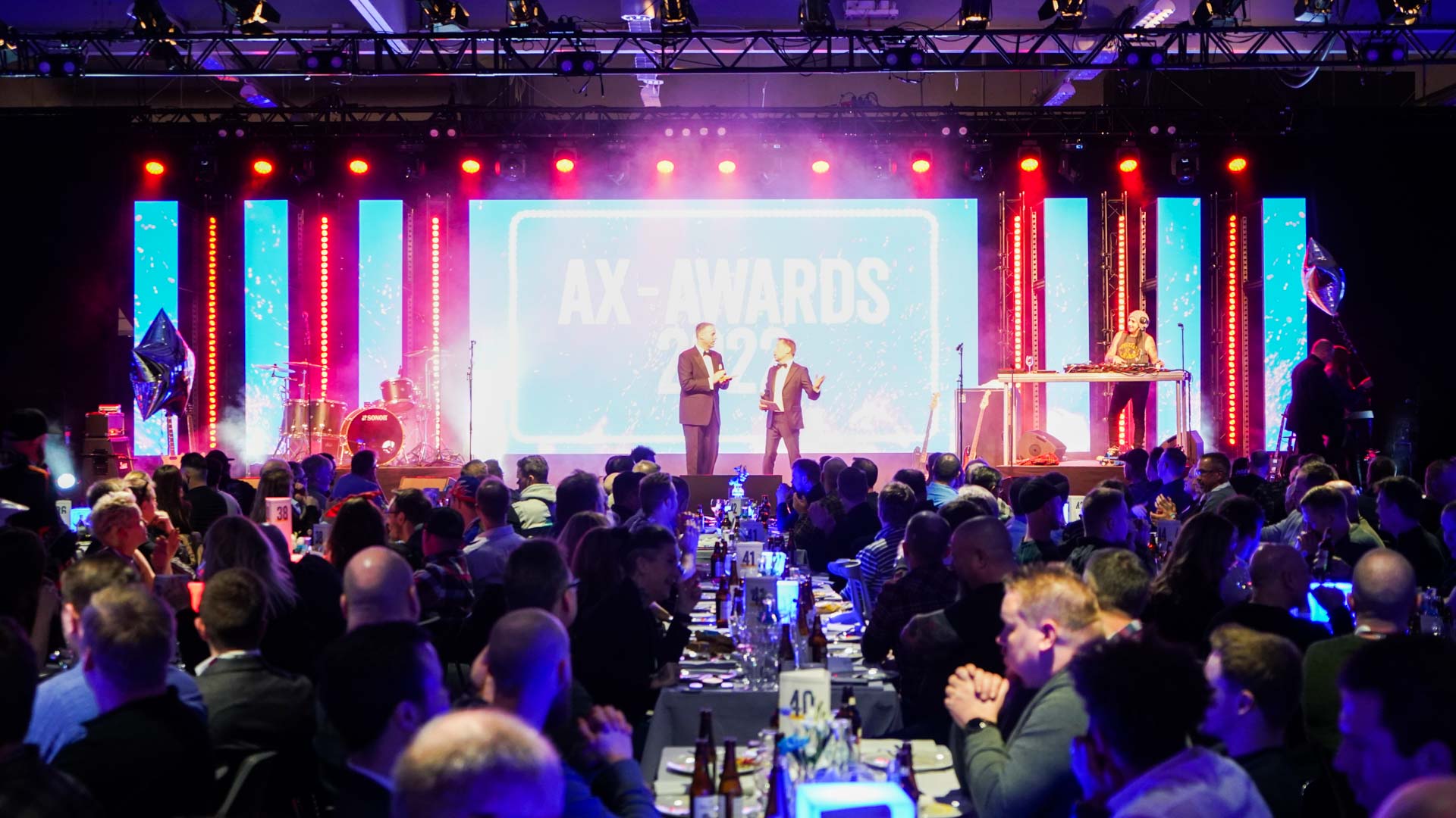 A large audience sits at banquet tables facing a brightly lit stage with two presenters standing in front of a big screen that reads AX AWARDS. Red and blue lights illuminate the event space.