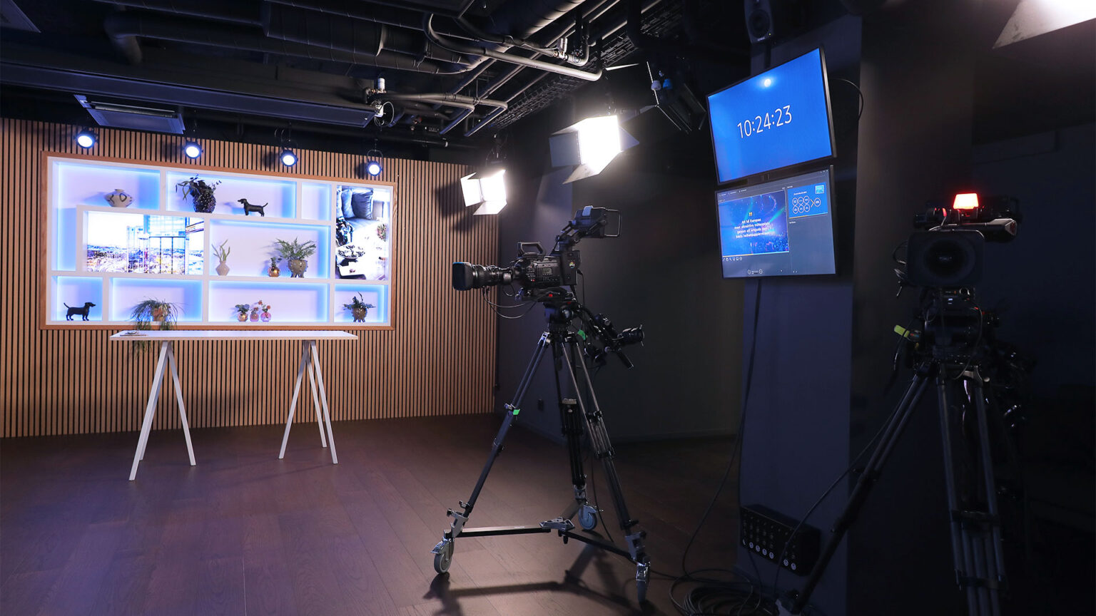 A modern TV studio with cameras on tripods, studio lights, a standing desk, shelves with decor and plants, and screens displaying time and information. The room has dark floors and a wood-paneled wall.