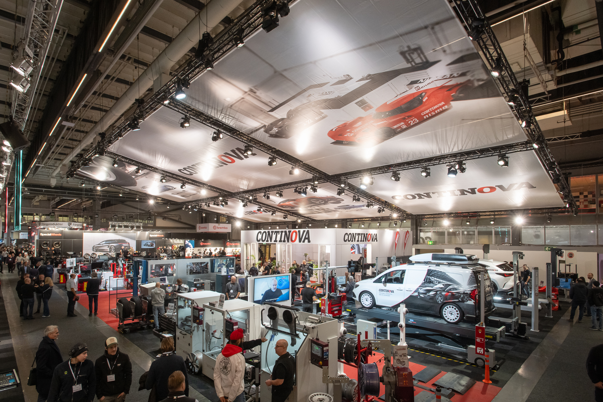 A busy automotive trade show booth features car lifts, equipment, and a white van on display, with many people walking around under a ceiling banner displaying cars and the CONTINOVA logo.