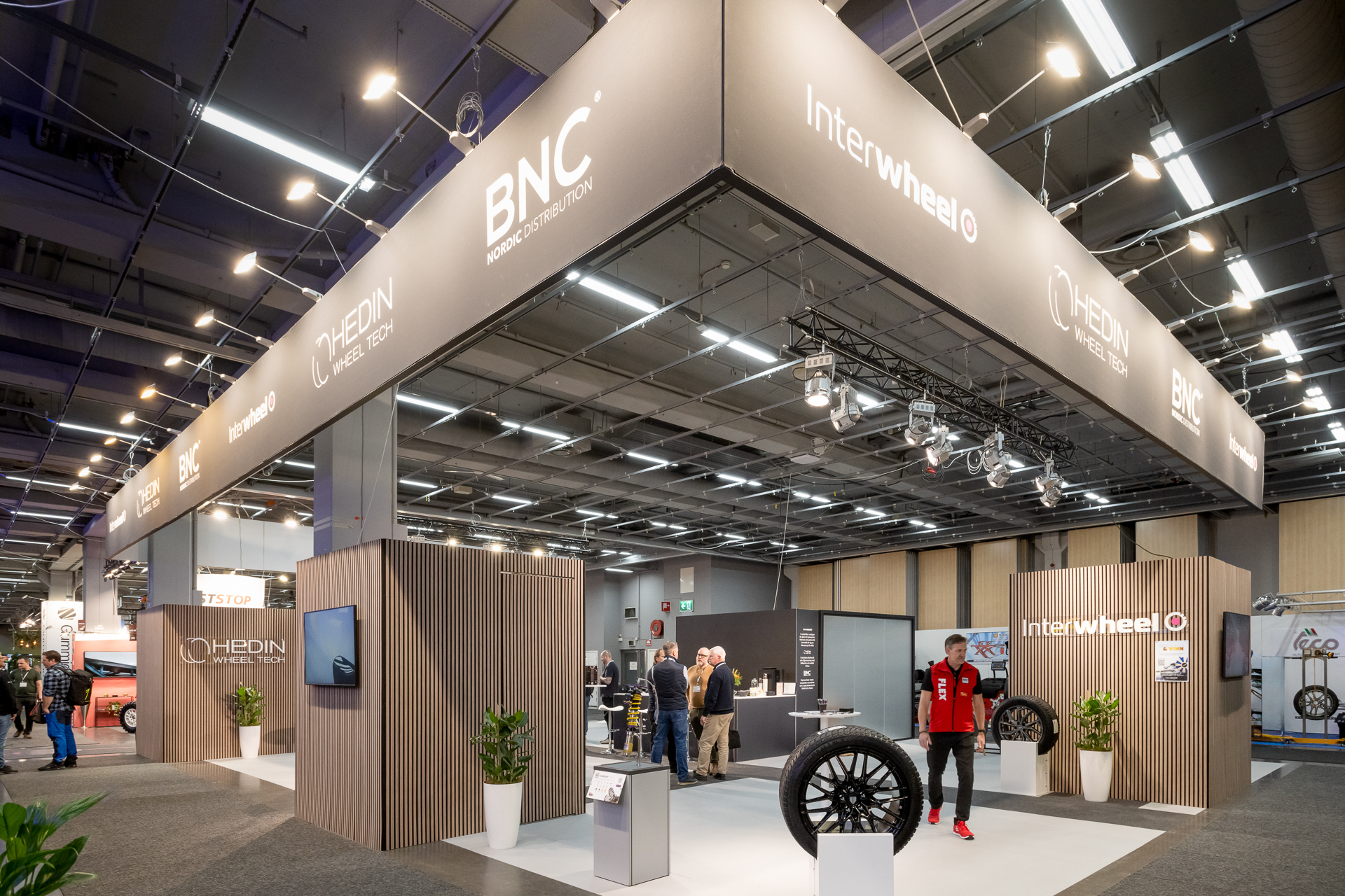 A modern exhibition booth with BNC and Interwheel logos, featuring sleek wood paneling, large banners, tires on display, potted plants, and people interacting in a bright, spacious convention hall.