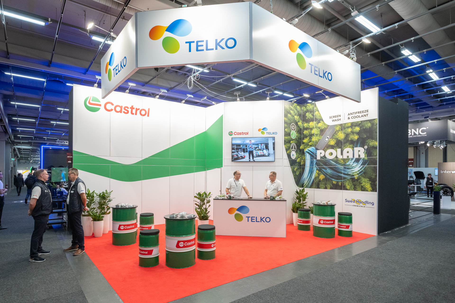 Trade show booth for Telko and Castrol, featuring branded signage, green barrels, potted plants, two staff at a counter, and display screens. Two attendees are conversing on the left side of the booth.