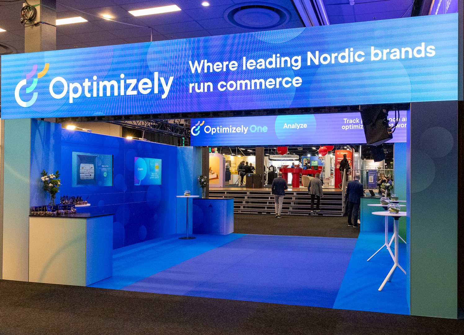 A trade show booth with a large digital sign reading “Optimizely: Where leading Nordic brands run commerce.” People walk inside, and the booth features blue décor, tables with flowers, and a modern, tech-focused atmosphere.