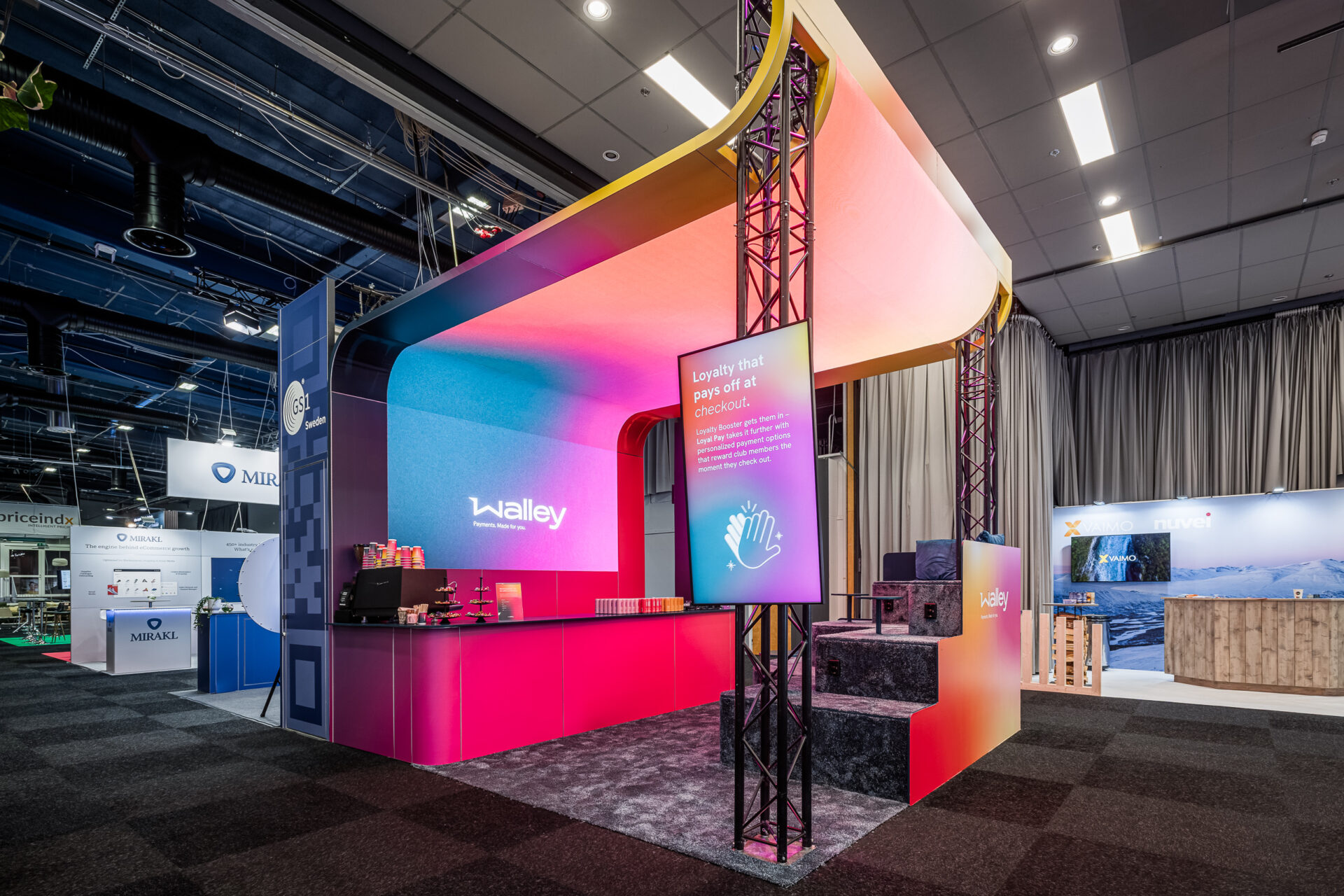A colorful exhibition booth with a curved, gradient canopy displaying the Walley logo, digital screens, counters, and product displays, set in a large, modern convention hall with other booths in the background.