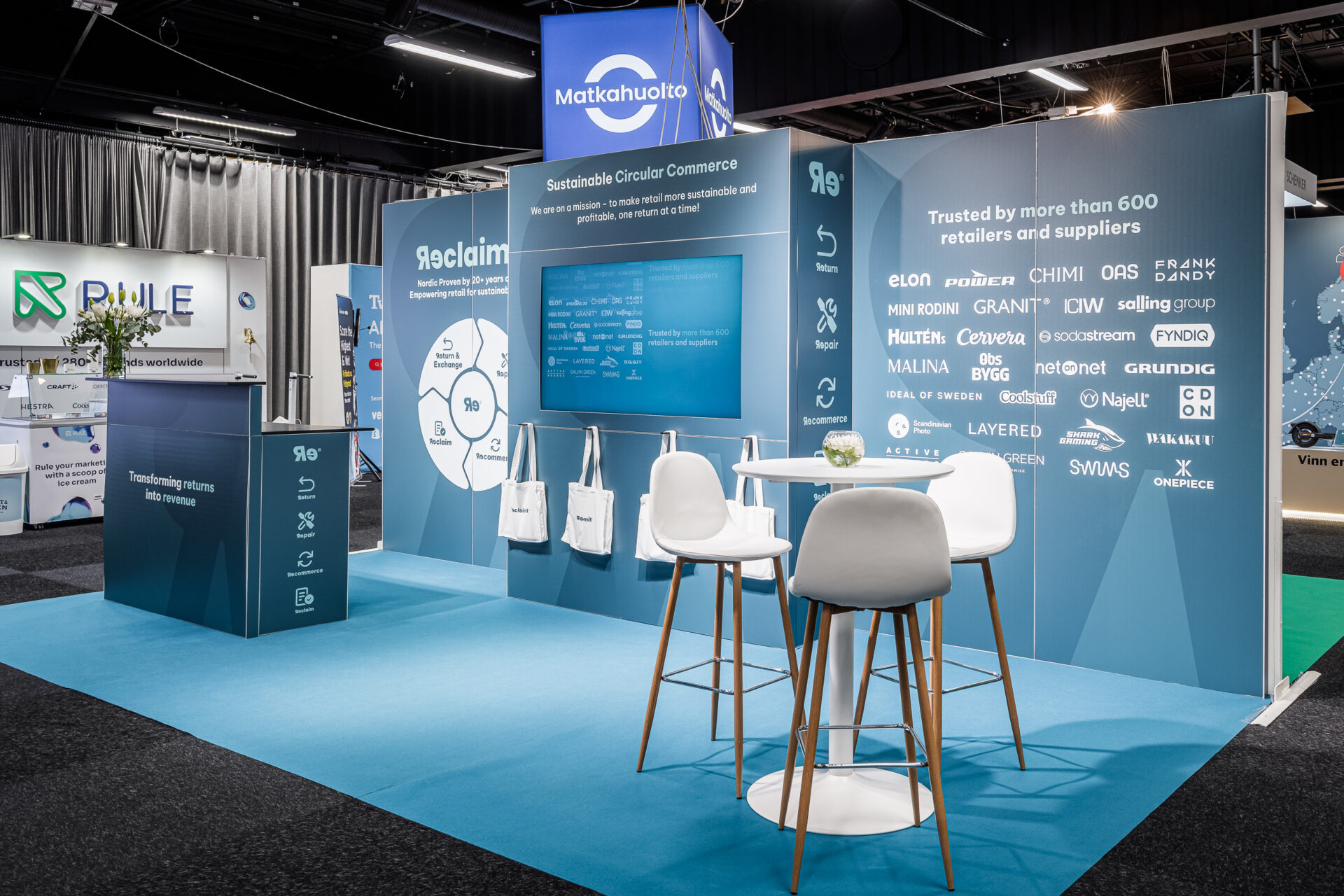 A modern exhibition booth with blue and white decor displays company names and information about sustainable circular commerce. The setup includes a screen, diagrams, high tables, and white chairs on a blue carpet.