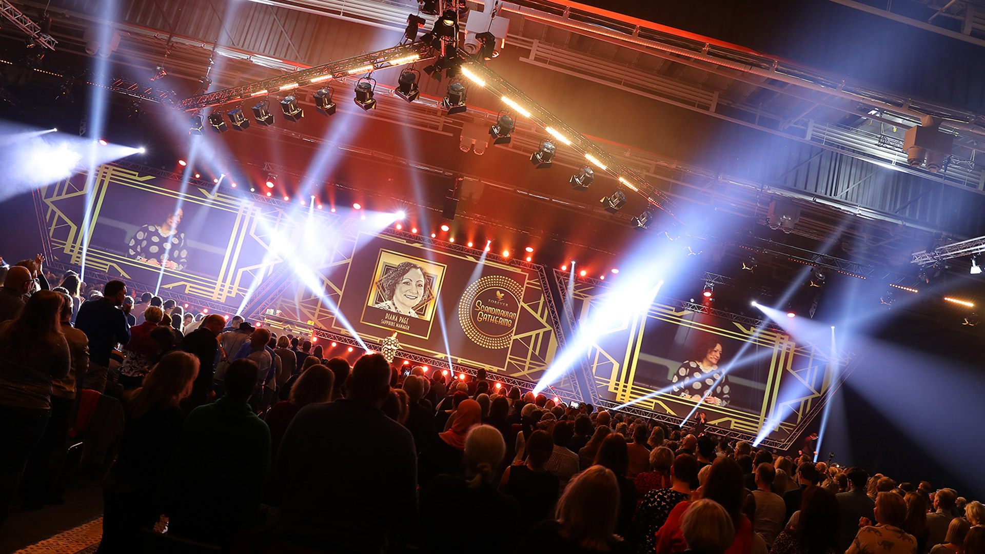 A large crowd stands facing a brightly lit stage with spotlights, digital screens displaying a woman’s portrait, and gold geometric designs, suggesting an award ceremony or celebration event in a spacious indoor venue.