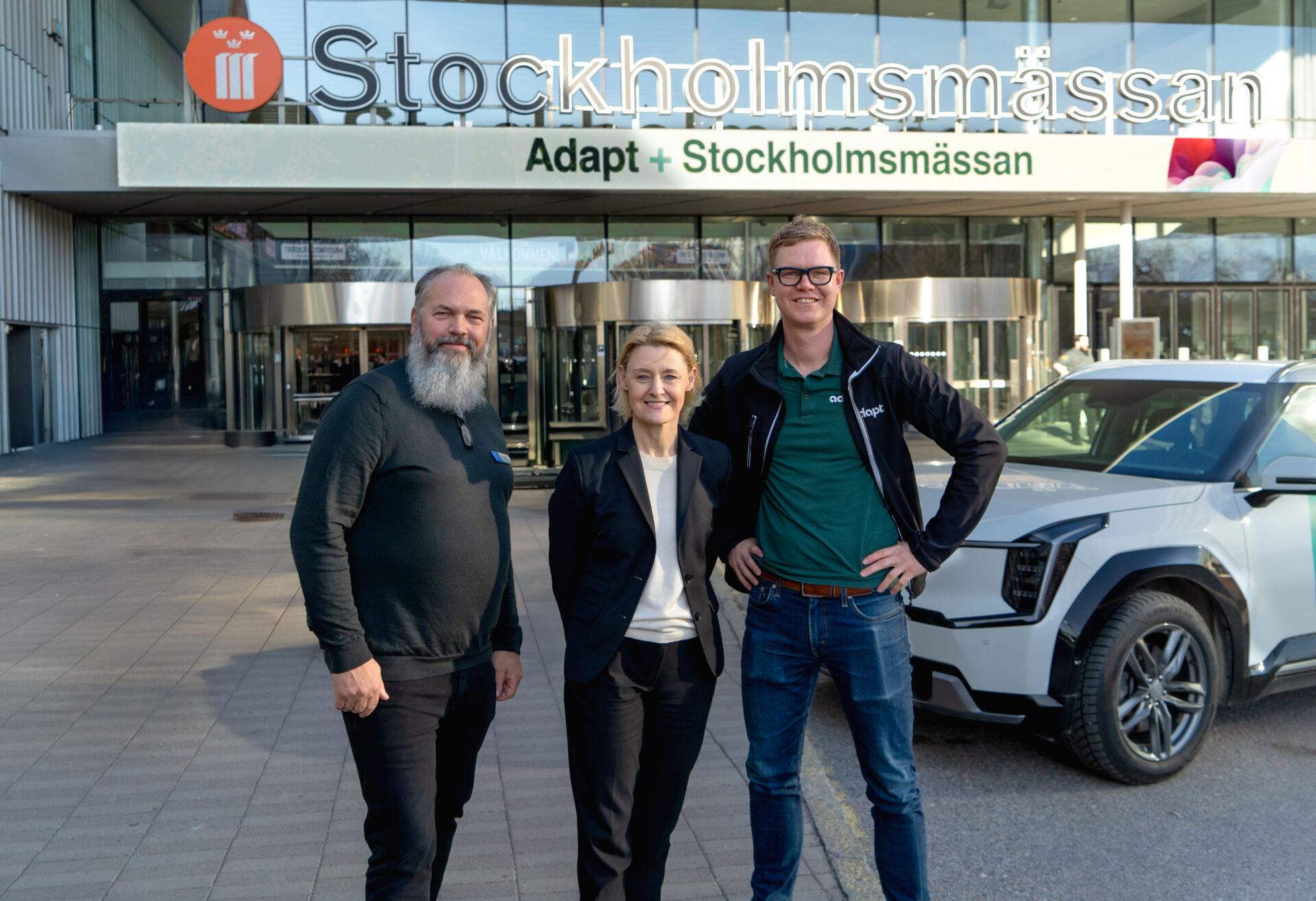 Three people stand smiling outside Stockholmsmässan convention center in Stockholm, with a white car parked nearby and the building’s signage visible in the background.