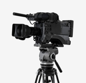 A professional video camera with a large lens and microphone mounted on a sturdy tripod, set against a plain white background.