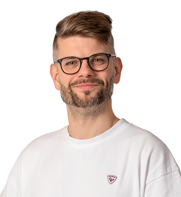 A man with short, light brown hair styled to the side, a trimmed beard, and glasses, wearing a white t-shirt with a small logo, smiling against a plain white background.