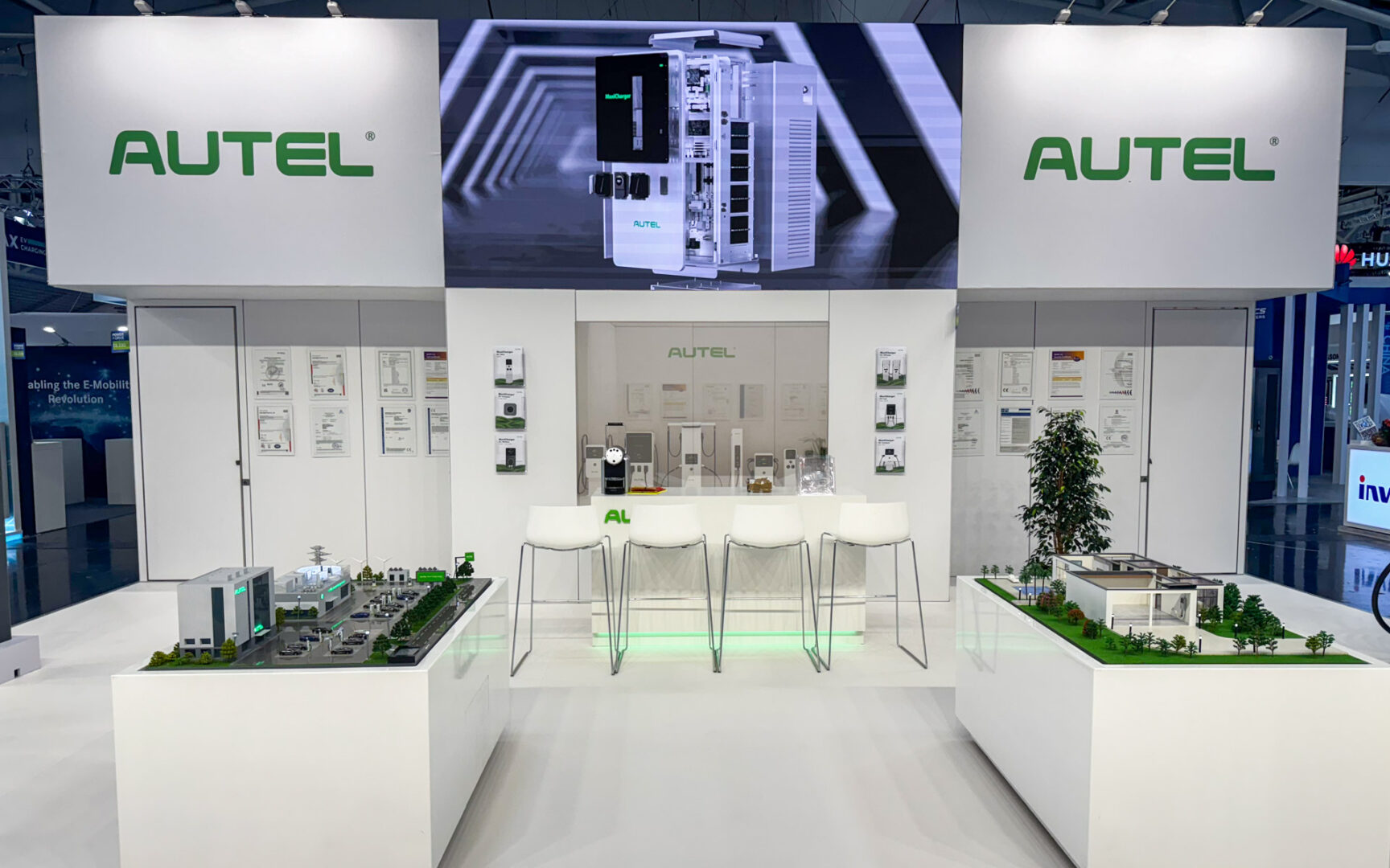 An exhibition booth for AUTEL features a white modern design, display panels, product models on tables, three white chairs, and a counter. Green AUTEL logos are prominently visible on walls and displays.