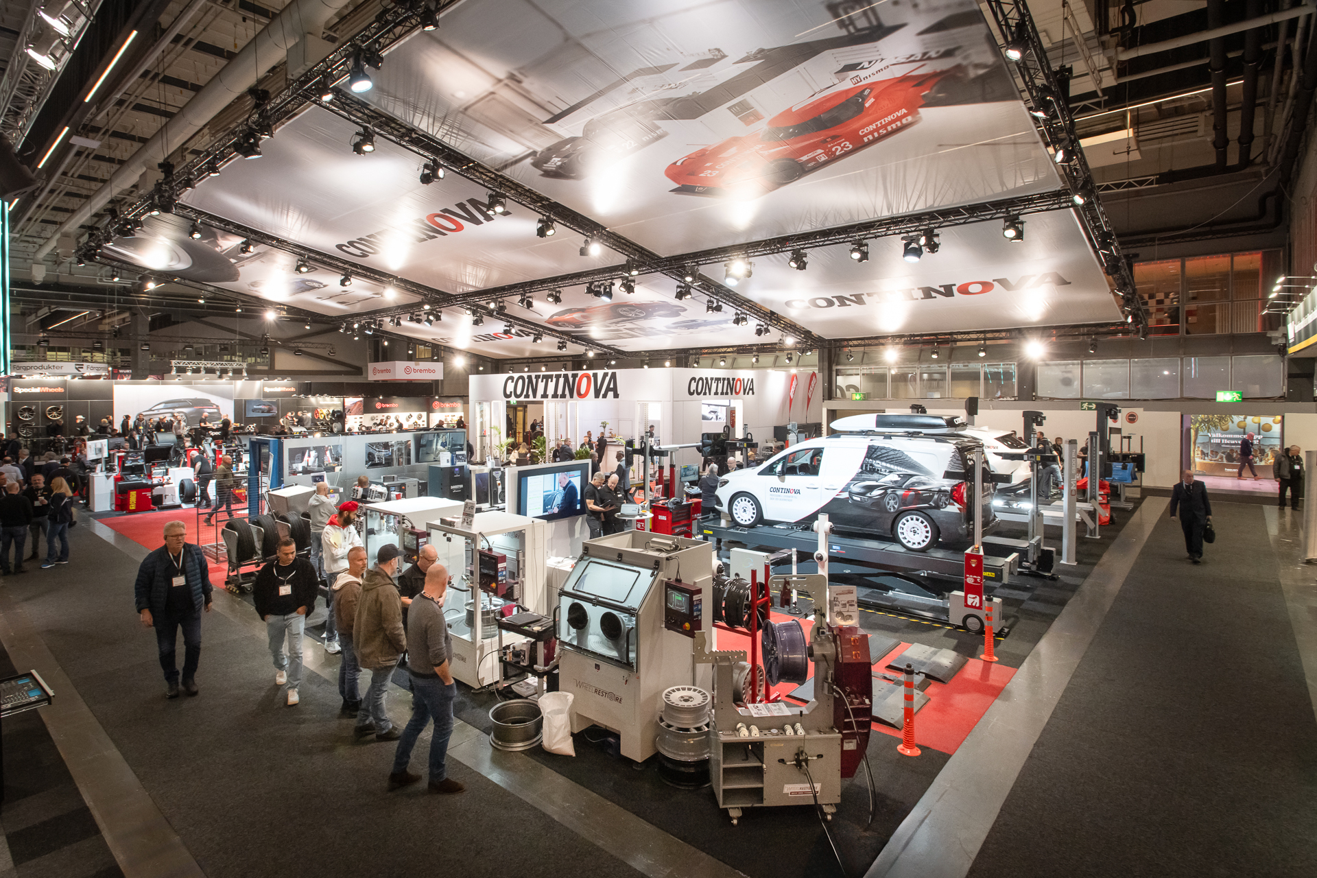 A busy trade show floor features multiple industrial equipment displays, visitors walking, and a prominent CONTINOVA booth. A white van and large machines are showcased under bright lighting in a spacious exhibition hall.