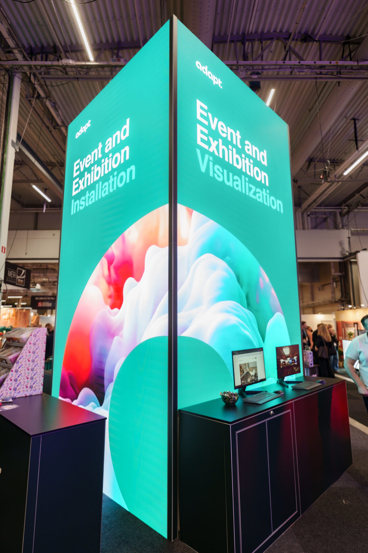 A tall, brightly lit exhibition booth features the text Event and Exhibition Installation and Event and Exhibition Visualization with colorful abstract graphics, computers on a black counter, and people in the background.