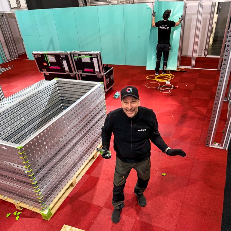 Two people assemble a room with teal wall panels and red carpet. One smiles at the camera, standing near stacked metal frames, while the other works with a wall panel in the background. Equipment and cables are scattered around.