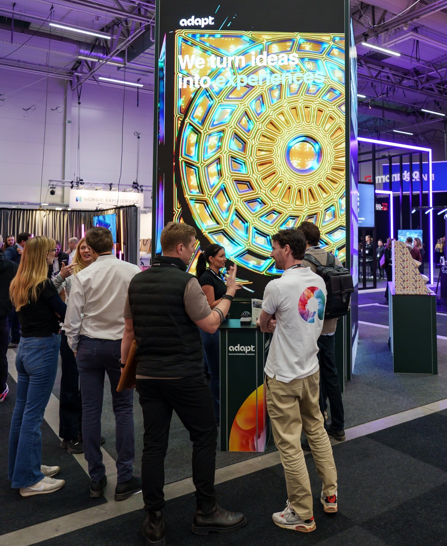 A group of people stand and talk in front of a tall digital display showing colorful geometric patterns and the words We turn ideas into experiences at an indoor event or exhibition.