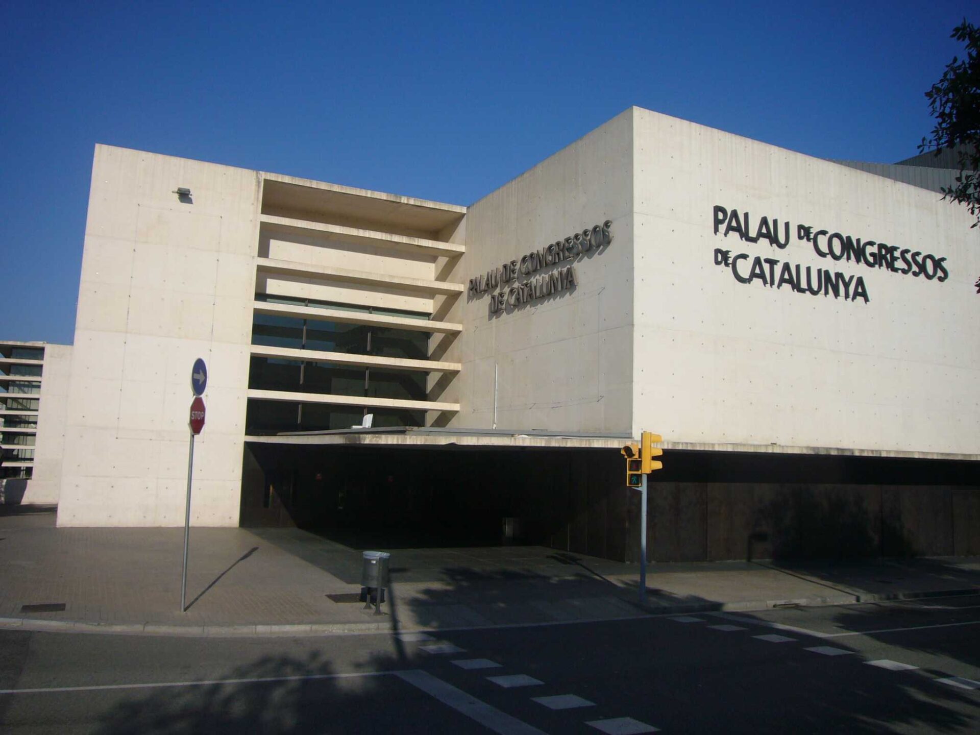 Modern white concrete building with large geometric shapes and the words Palau de Congressos de Catalunya on its facade, seen on a sunny day with a blue sky. A traffic signal and sidewalk are in the foreground.