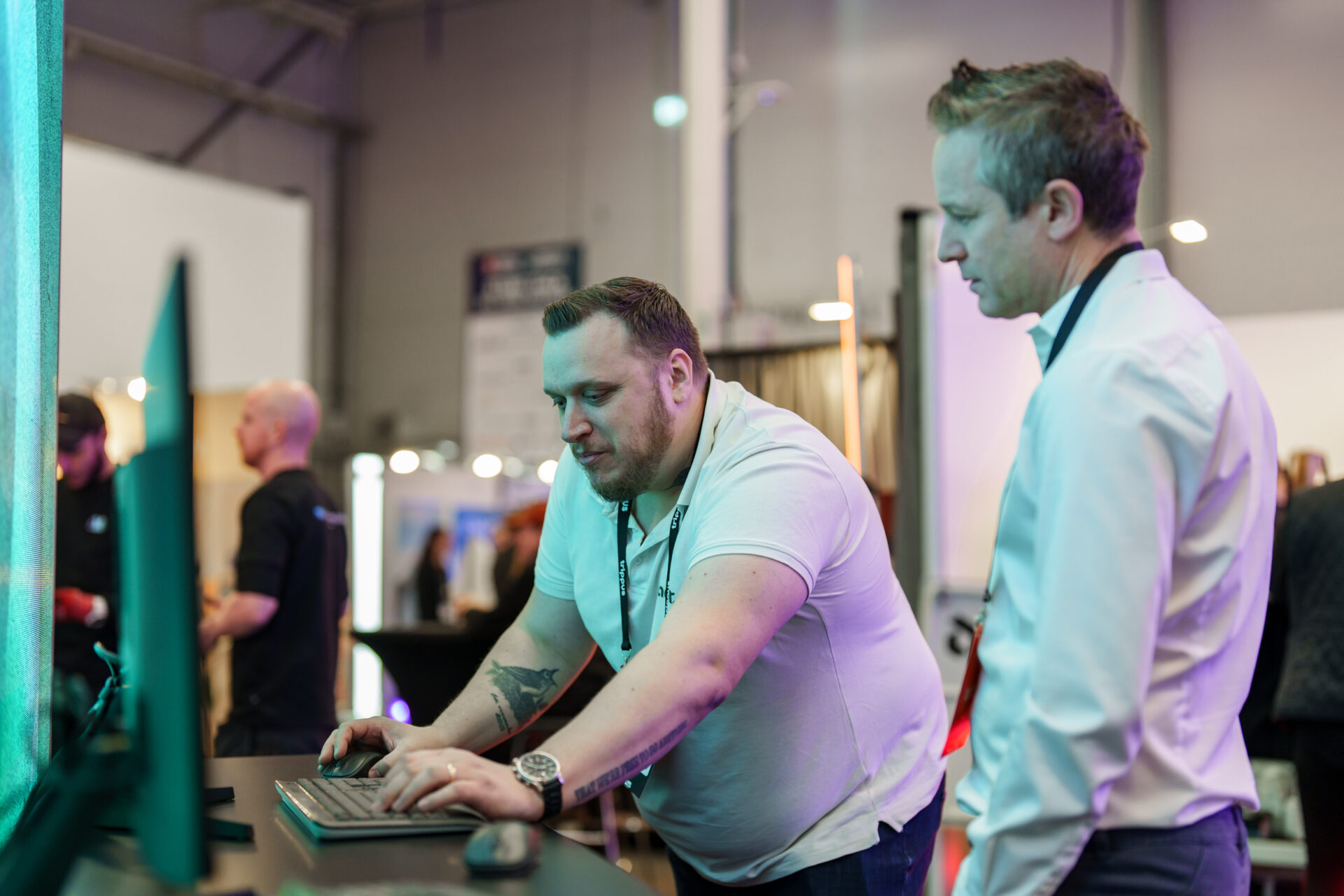 Two men stand at a desk with a computer; one is typing on the keyboard while the other looks at the screen. Both wear smart casual clothes at what appears to be a professional event or expo with others in the background.