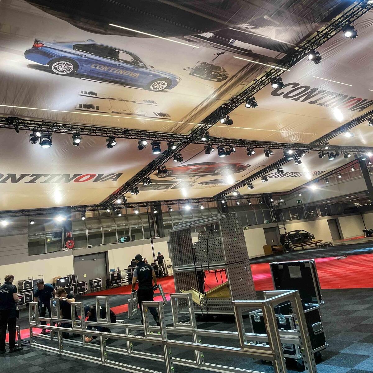 Large indoor event space being set up, with workers assembling equipment. The ceiling is covered by large banners featuring a blue car and the word “CONTINOVA.” Lighting rigs and red flooring are visible.