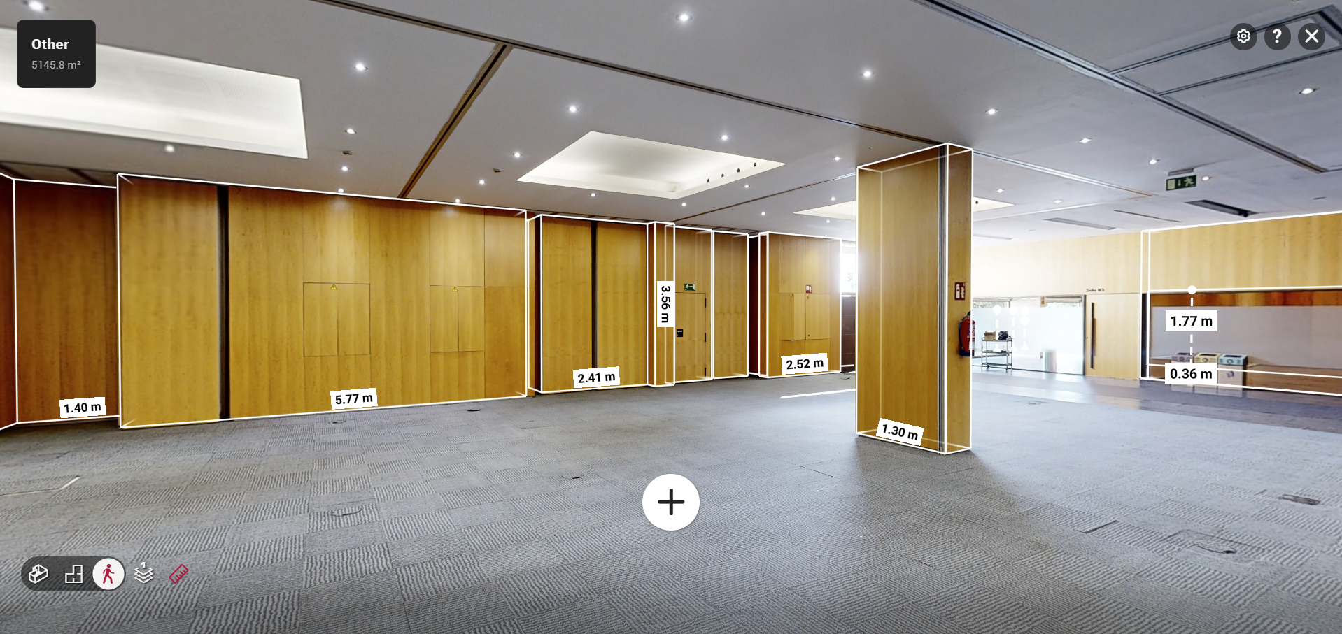A spacious, empty conference room with wooden partition walls and a gray carpet. Various wall and door measurements are marked in white text throughout the room. Ceiling lights and skylights provide illumination.