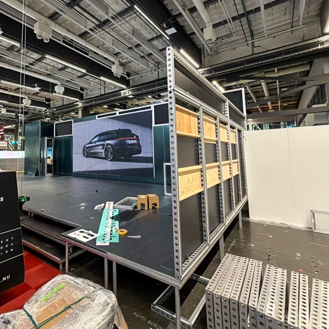 An indoor exhibition setup in progress with metal frames, wooden crates, and construction materials. A large digital screen displays a car image on the back wall. The area is industrial with exposed ceilings.