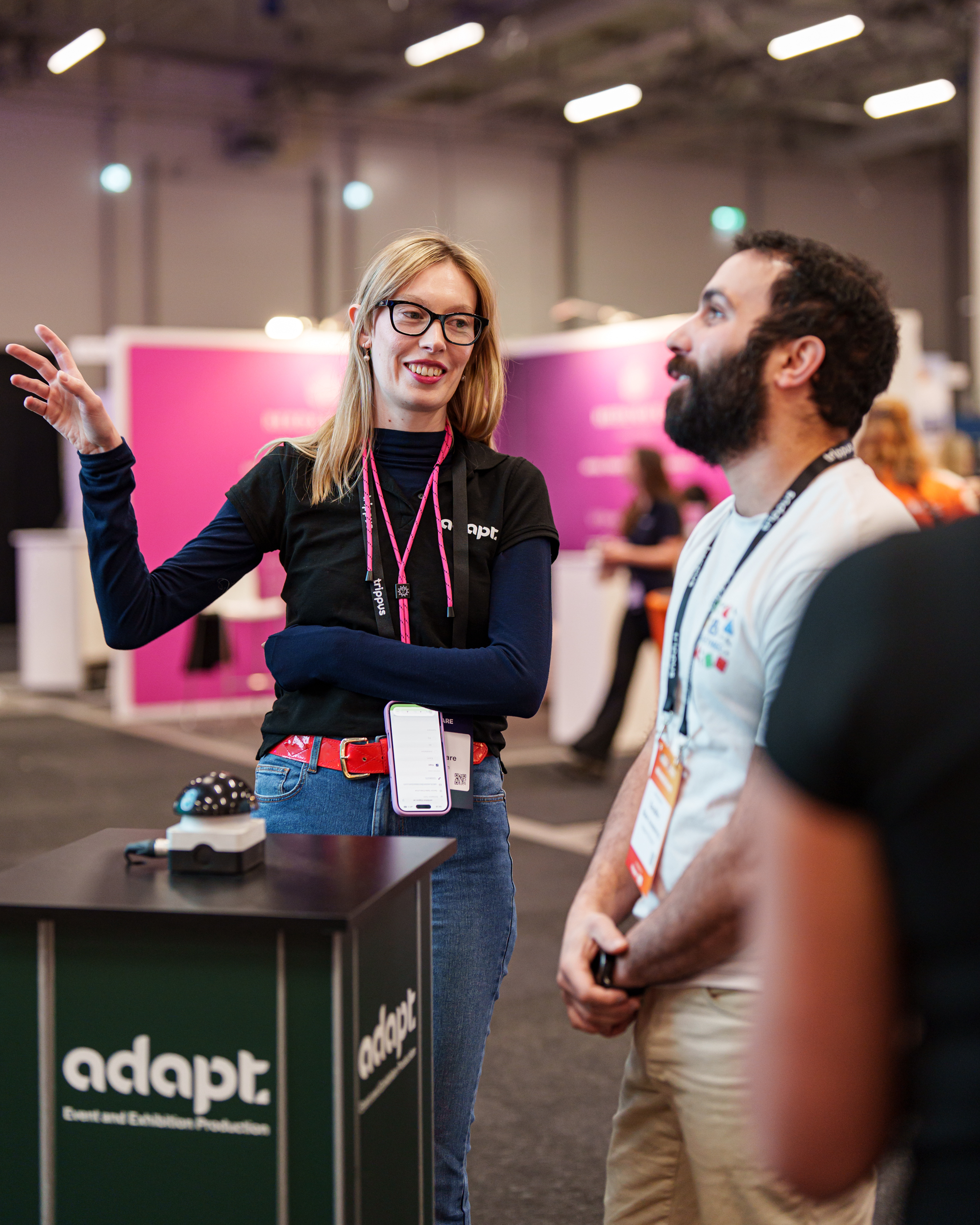 A woman and a man are standing and smiling in conversation at a conference. The woman gestures with her hand while standing beside a booth labeled “adapt.” Colorful booths and other people are visible in the background.