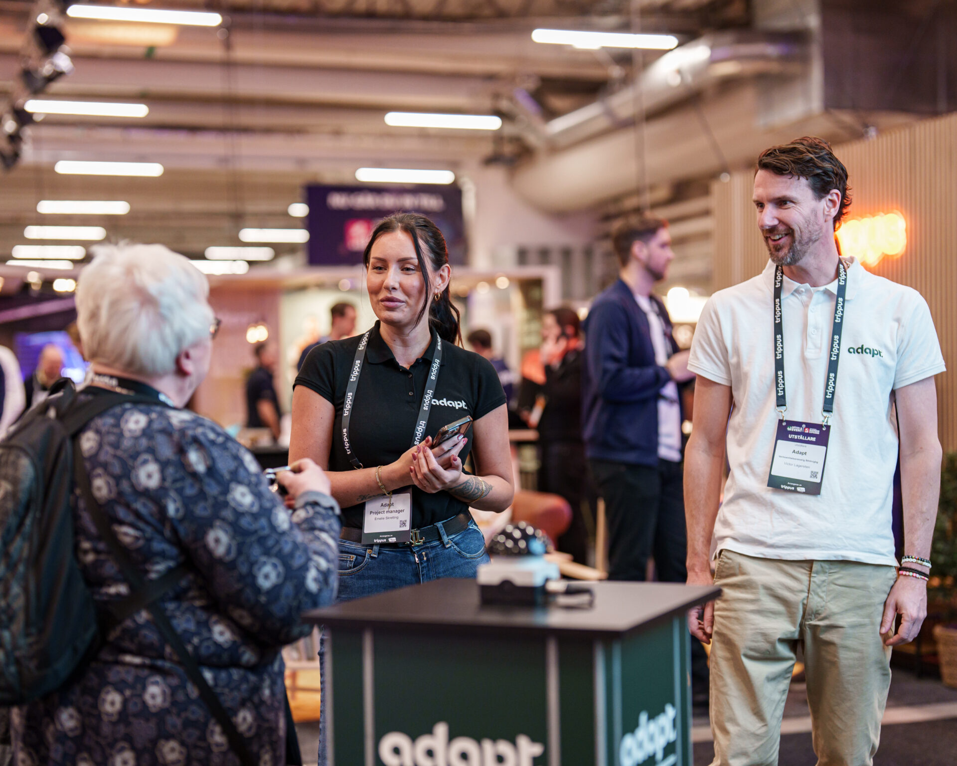 Three people are talking at an indoor event or expo. Two staff members with name badges stand behind a booth labeled adapt while an older person stands in front, engaging in conversation. The background is busy with other attendees.