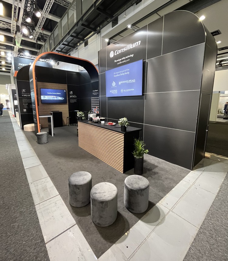A modern exhibition booth for Consillium with a sleek black design, digital screens, a reception counter, small potted plants, and several gray velvet stools arranged in front.