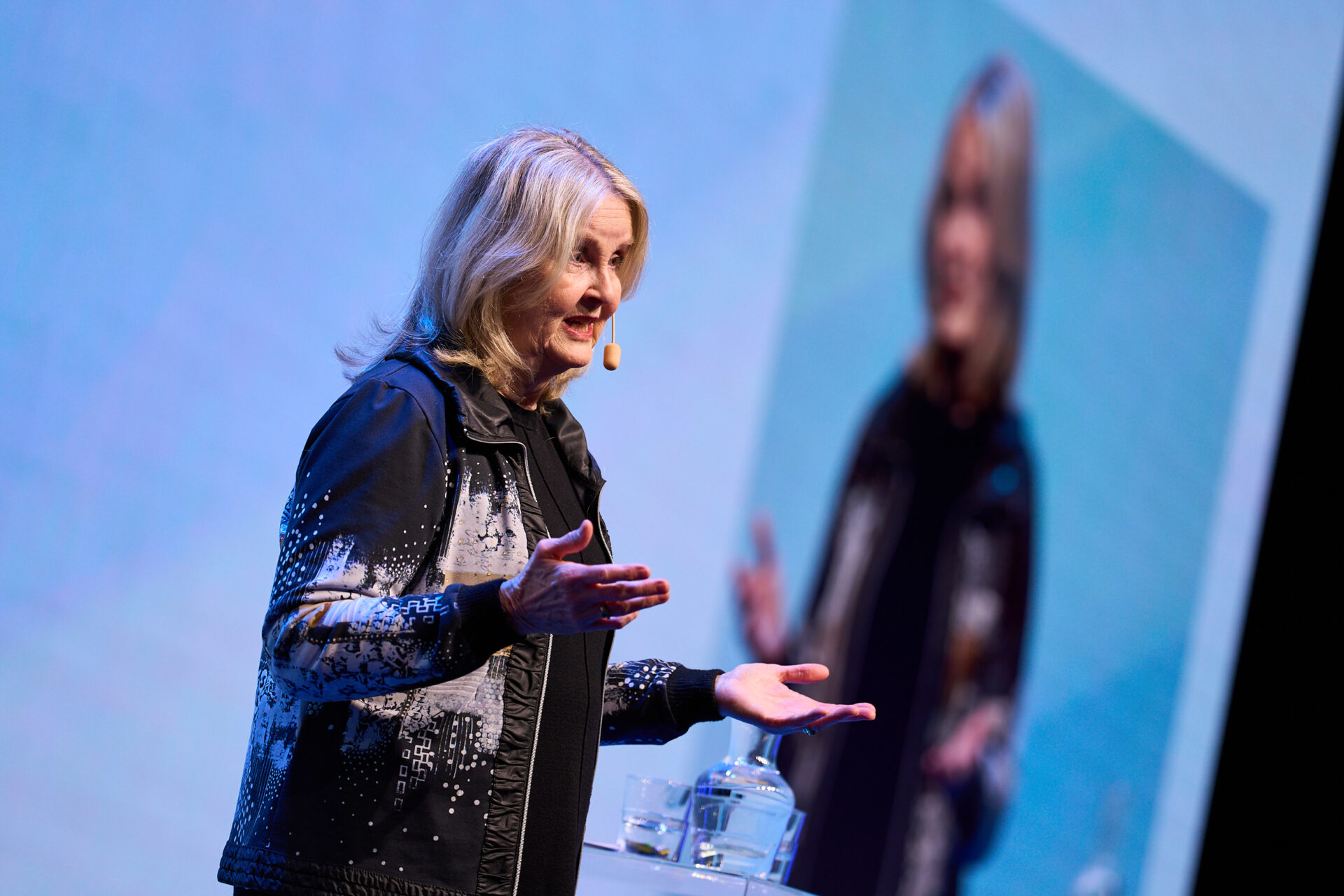 A woman with blonde hair wearing a black and white jacket speaks on stage with a headset microphone, gesturing with her hands. Her reflection is visible on the large screen behind her.