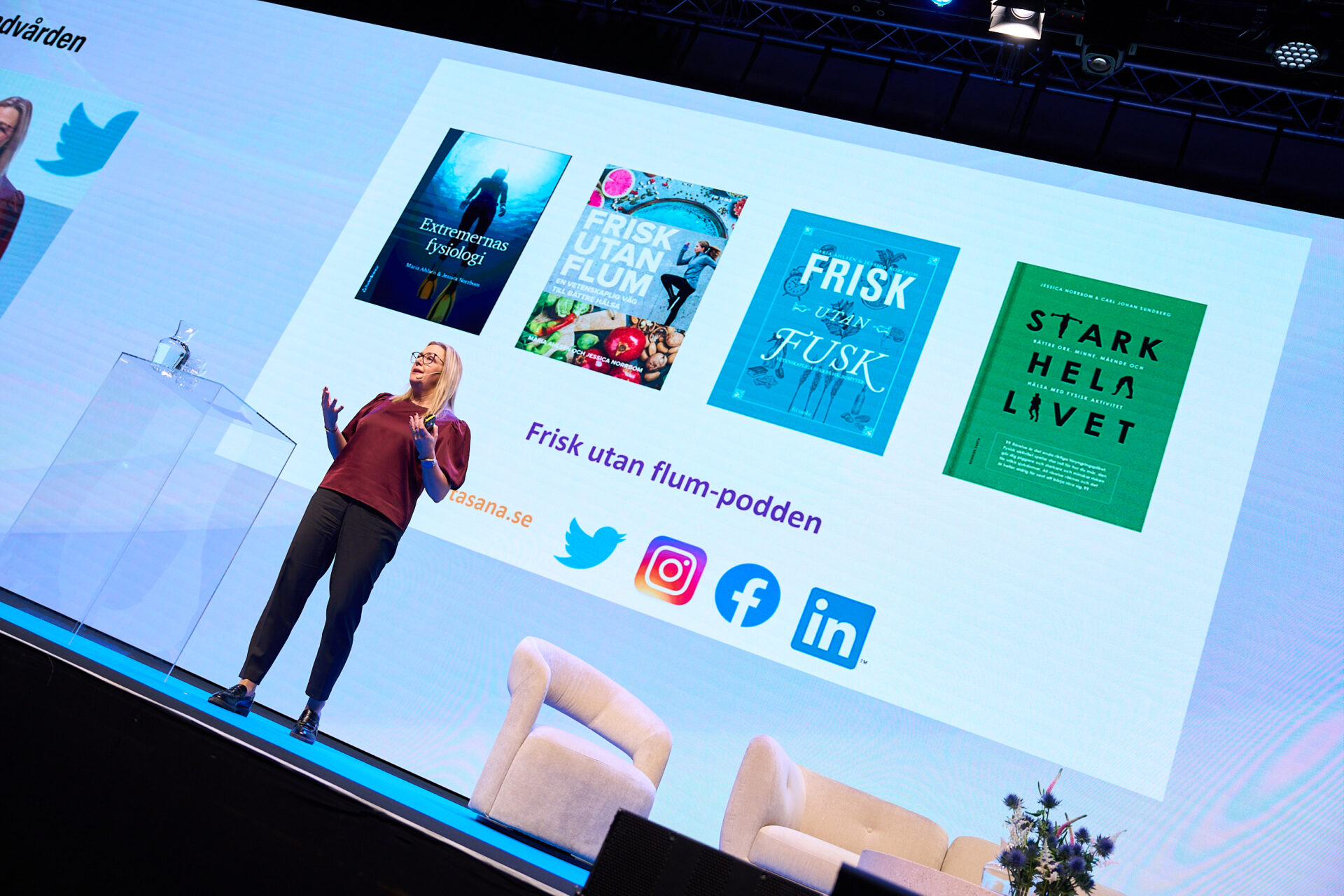 A woman speaks on stage in front of a screen displaying four book covers, podcast information, and social media icons for Twitter, Instagram, Facebook, and LinkedIn at a professional event.