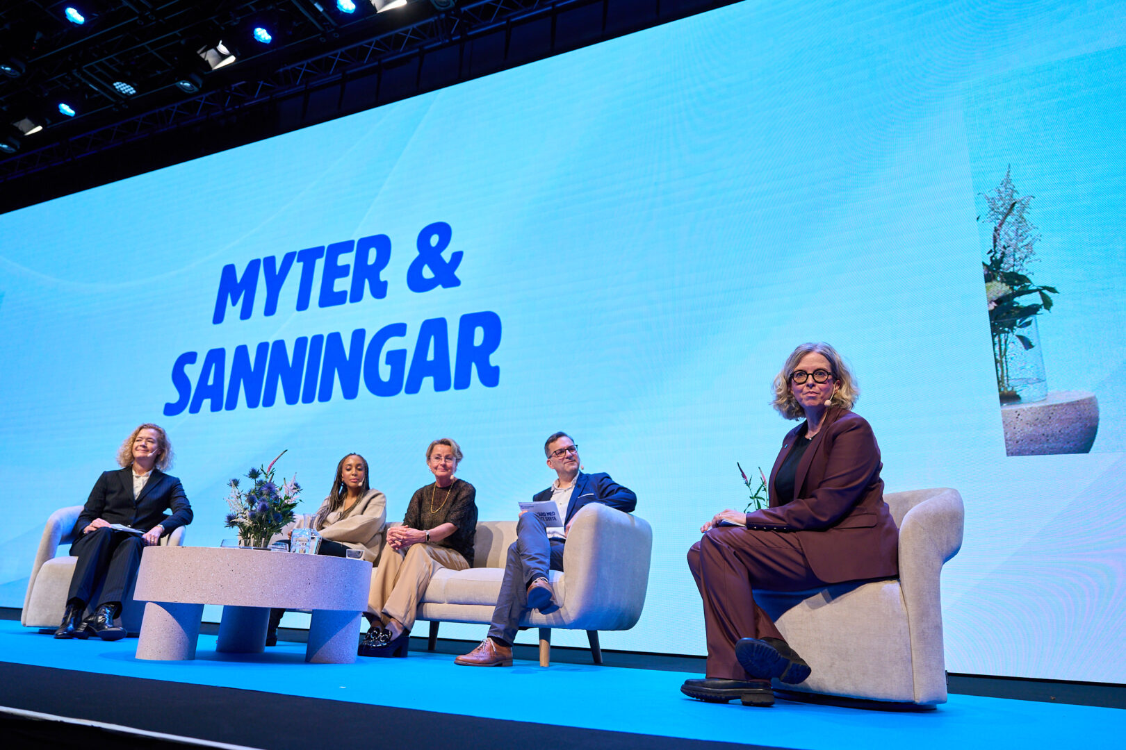 Five people sit on stage in armchairs under a large screen displaying the Swedish words MYTER & SANNINGAR. The panelists are dressed in business attire. A small table with flowers is in front of them.