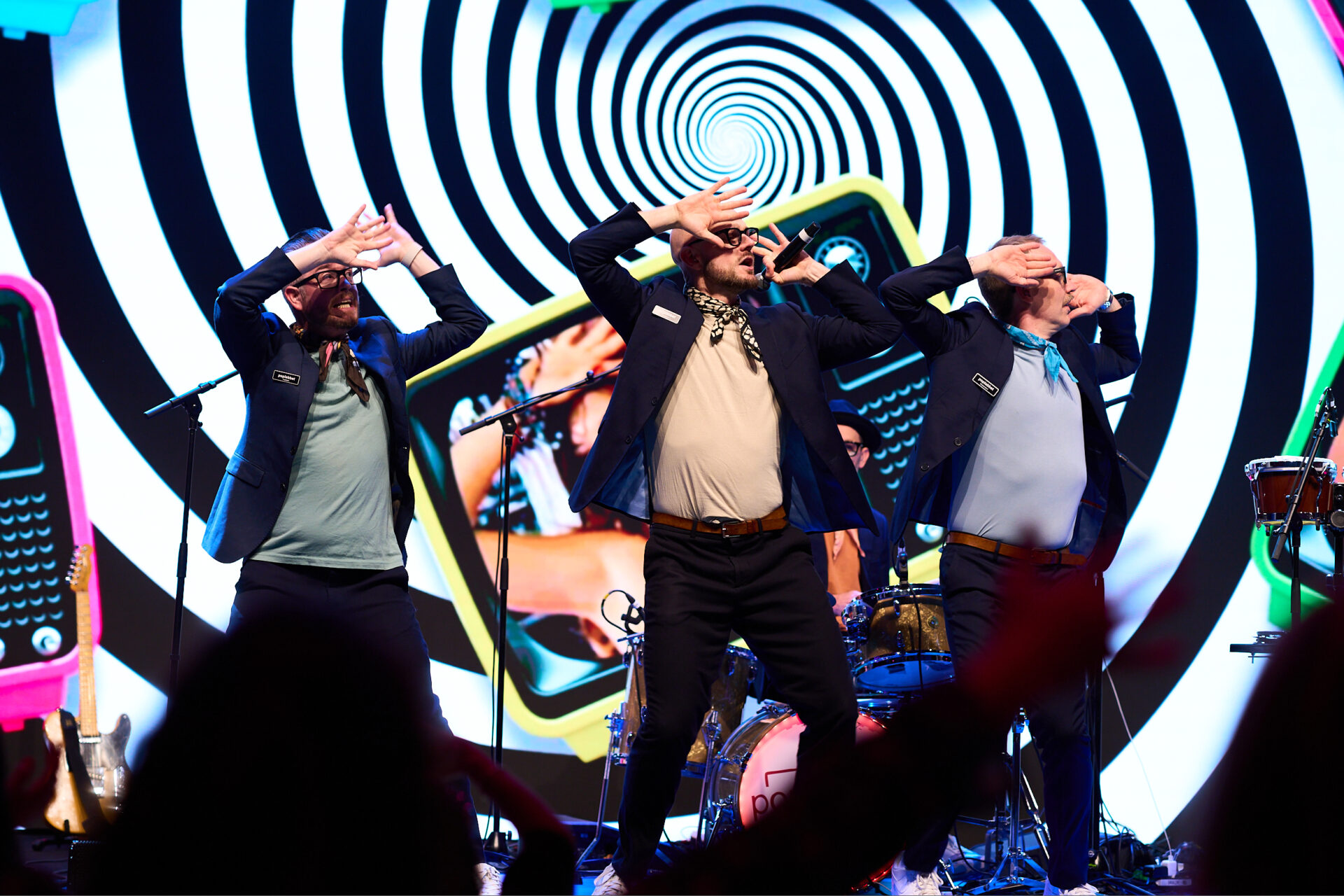 Three men in matching jackets perform energetically on stage with their hands on their foreheads. Behind them is a colorful, hypnotic spiral backdrop featuring cartoonish phones. Musical instruments and audience silhouettes are visible.