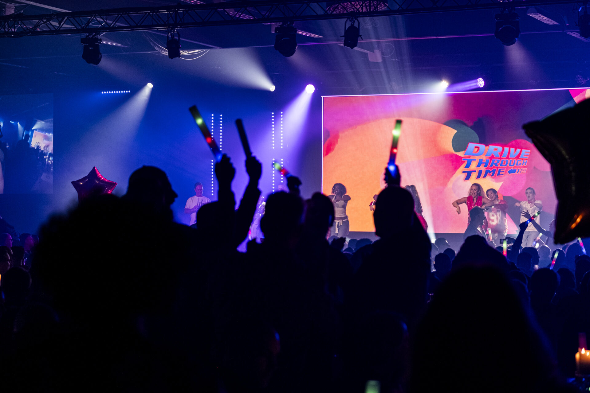 A crowd waves glow sticks at a lively indoor event with a colorful stage, bright lights, and a large screen displaying Drive Through Time. Performers are visible on the stage.