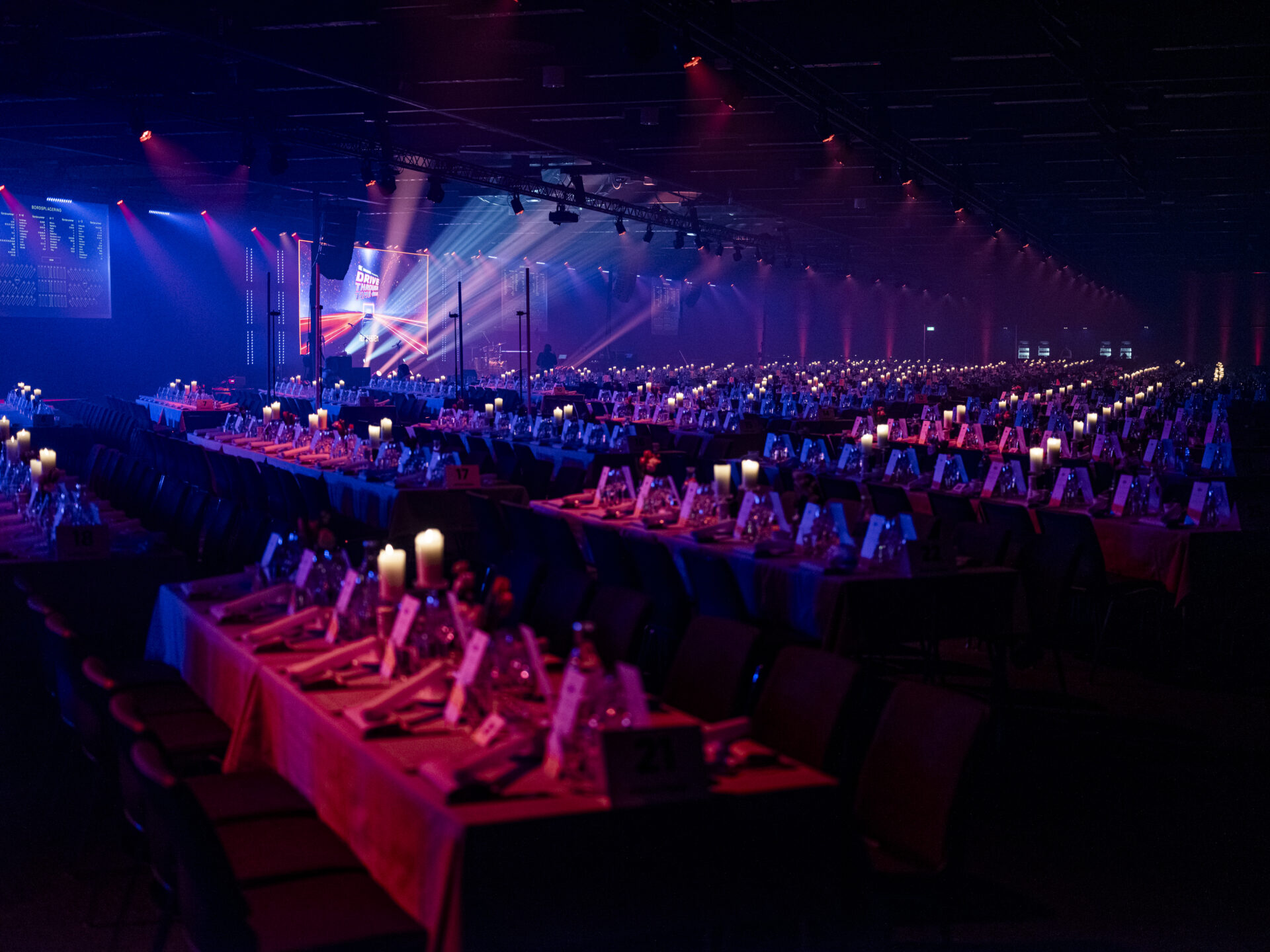 A large, dimly lit banquet hall with rows of long tables set for an event. Tables are decorated with candles and place settings, and spotlights cast colorful beams across the room.