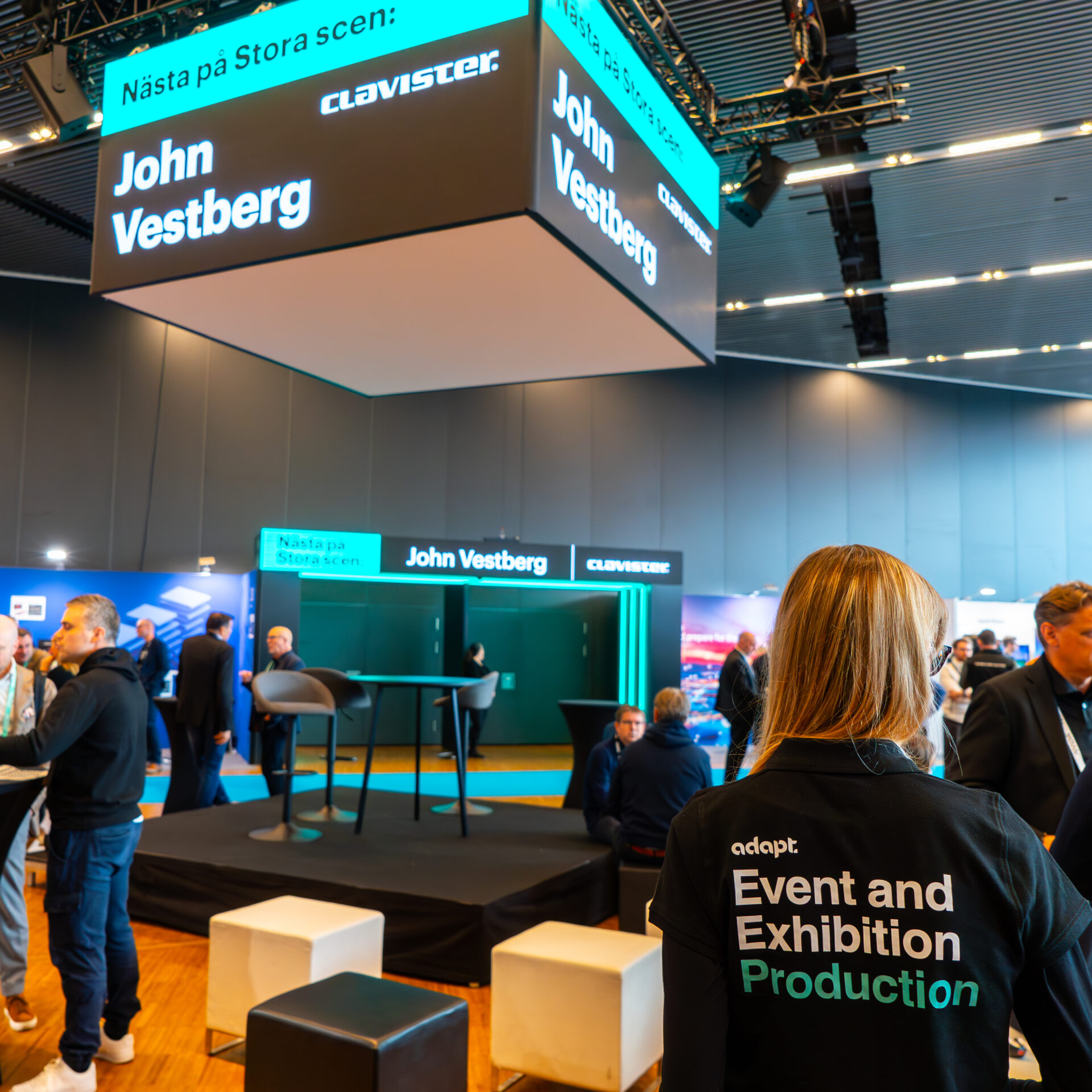 A woman wearing an Event and Exhibition Production jacket stands in a modern conference hall. Overhead screens display “John Vestberg” and “Clavister.” Attendees are gathered around, engaging and networking.