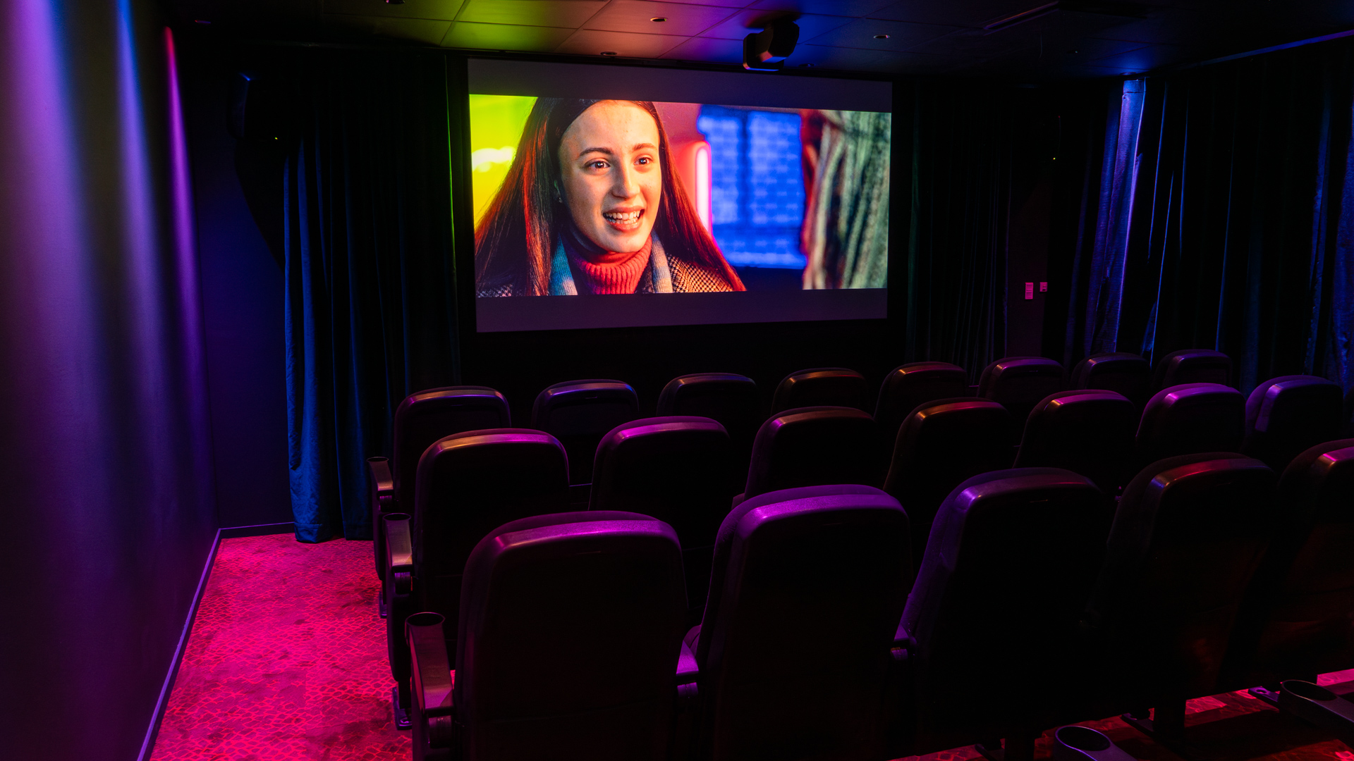 A small, empty movie theater with rows of black seats faces a large screen showing a woman smiling. The room is lit with colorful purple and blue accent lights.