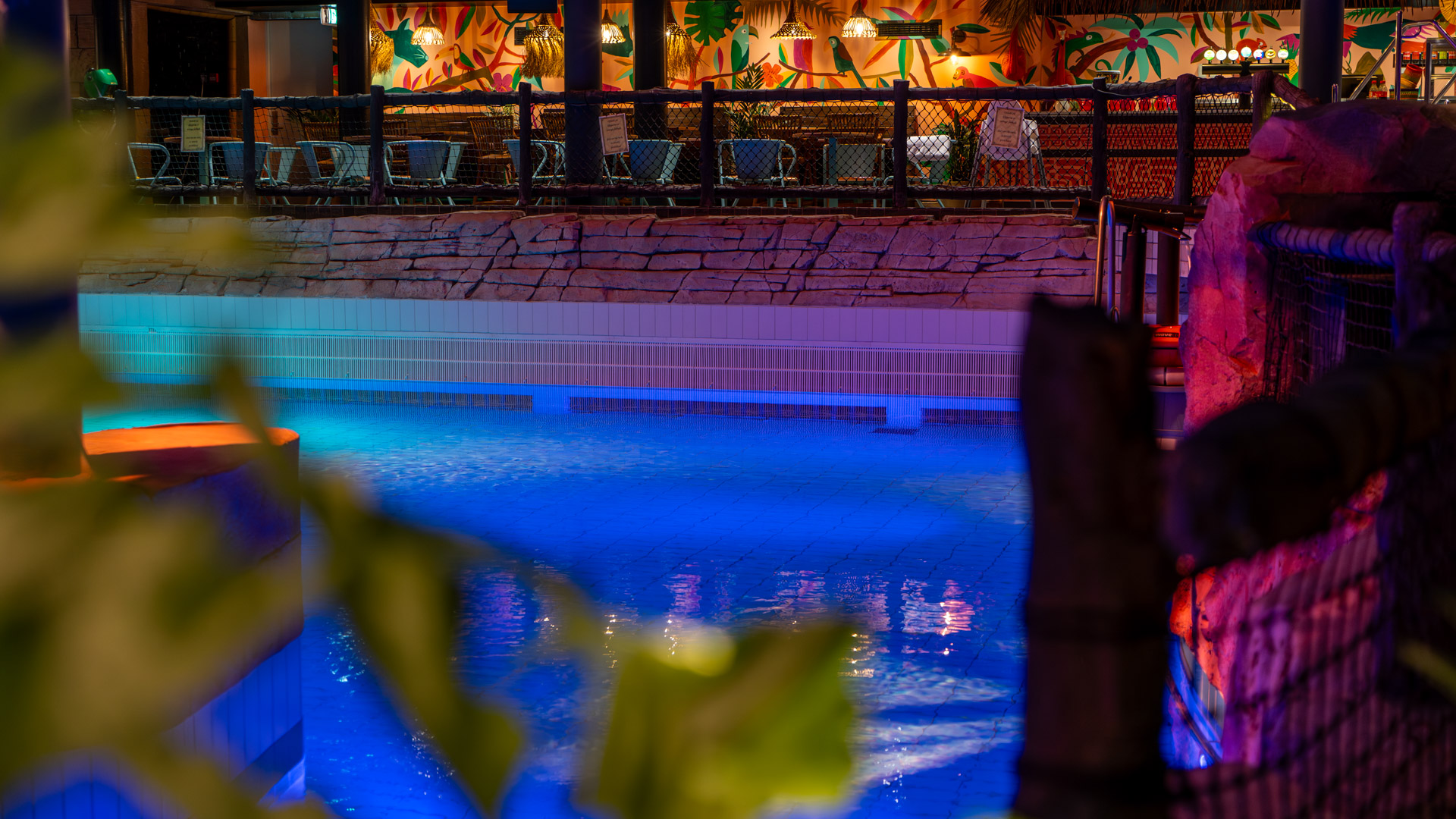 A brightly lit indoor pool glows blue at night, bordered by artificial rocks and fencing. In the background, empty tables and chairs sit beneath colorful, tropical-themed wall art.