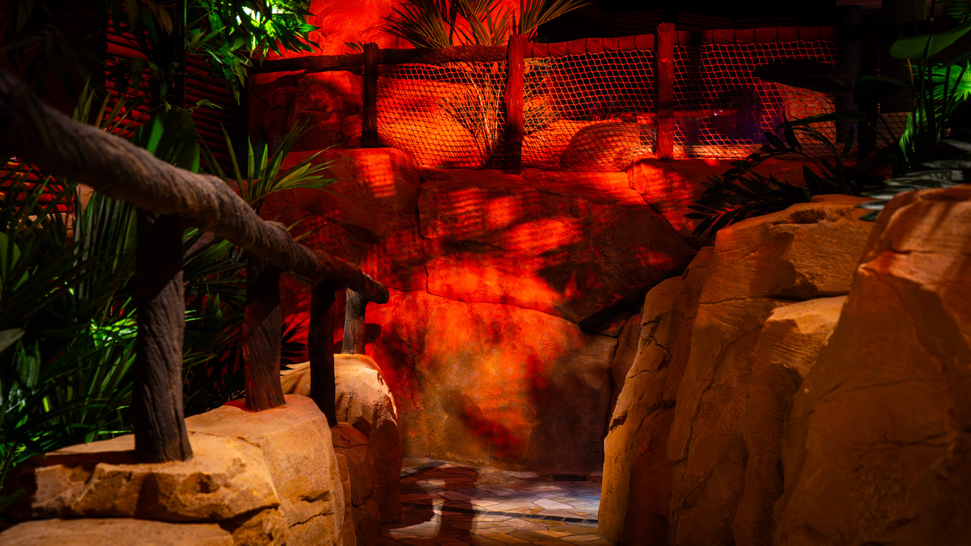 A pathway with a wooden railing runs between large artificial rocks, tropical plants, and is illuminated by warm, red-orange lighting, creating a jungle-like atmosphere.