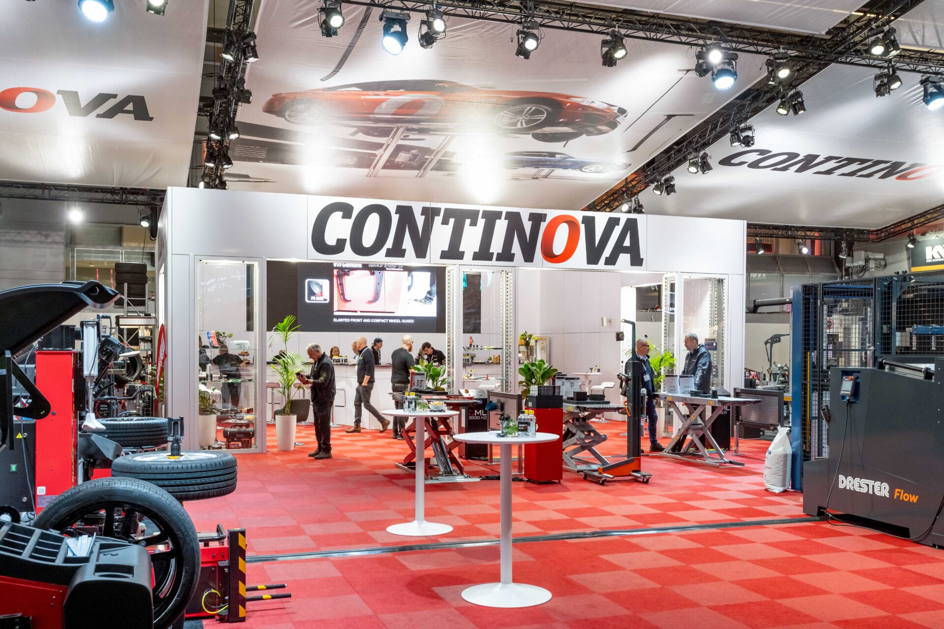 A trade show booth for CONTINOVA with a white and orange sign, surrounded by automotive equipment and machinery on a red checkered floor. People are interacting inside the booth under bright lights.