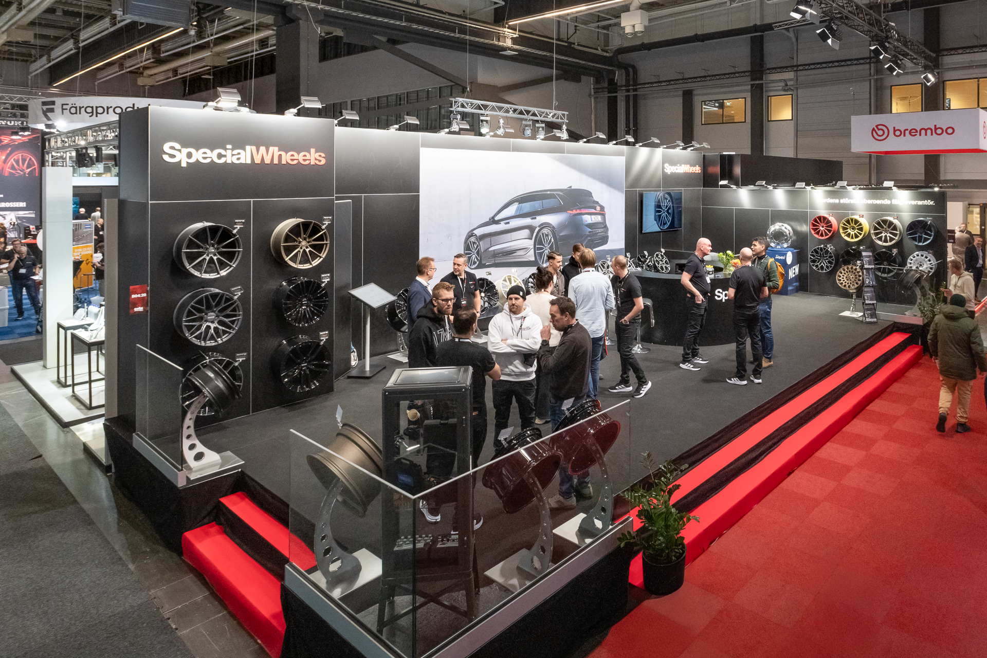 A group of people gathers at a car wheel exhibition booth labeled SpecialWheels, featuring various car rims on display. The booth is brightly lit, and there are other exhibitors in the background.