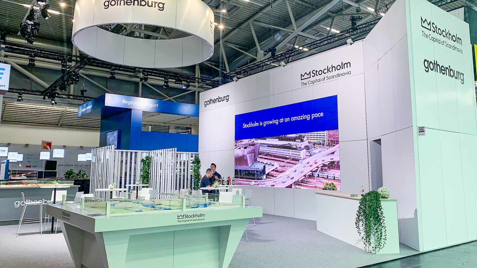 A modern exhibition booth for Stockholm and Gothenburg with a large digital screen displaying city images, architectural models on tables, and informational displays in a bright, spacious hall.
