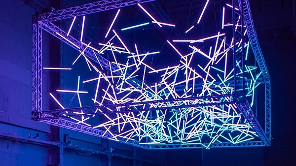 A suspended rectangular metal frame holds numerous glowing neon rods in blue, purple, and pink, creating an intricate, futuristic light installation against a dark background.