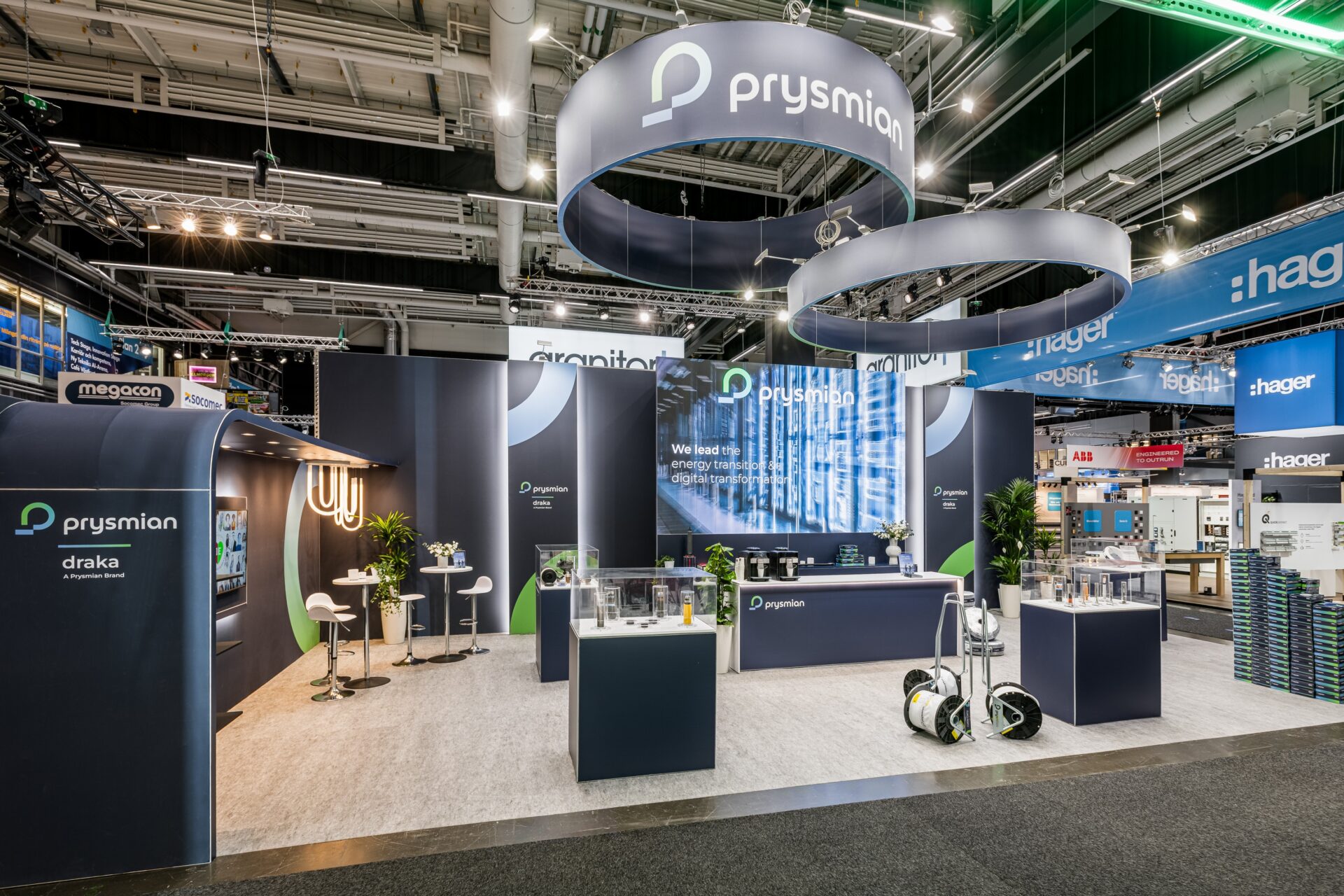 A modern exhibition booth for Prysmian features displays, seating areas, interactive screens, and hanging circular signs with the Prysmian logo, all set in a brightly lit convention hall.