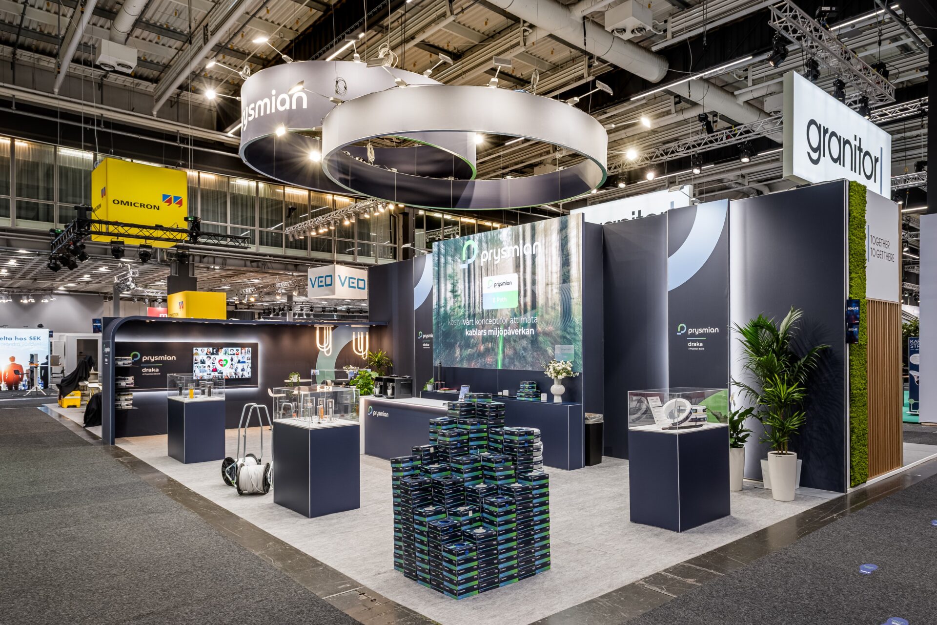 A modern exhibition booth with the name Prysmian displayed, featuring products on counters, digital screens, and promotional materials. The booth has contemporary decor, plants, and hanging circular lights in a large exhibition hall.