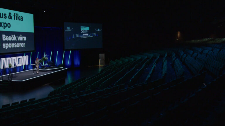 A speaker stands on a lit stage in a dark auditorium with rows of empty seats. A large screen above displays Swedish text and event information. Blue lights accent the stage area.